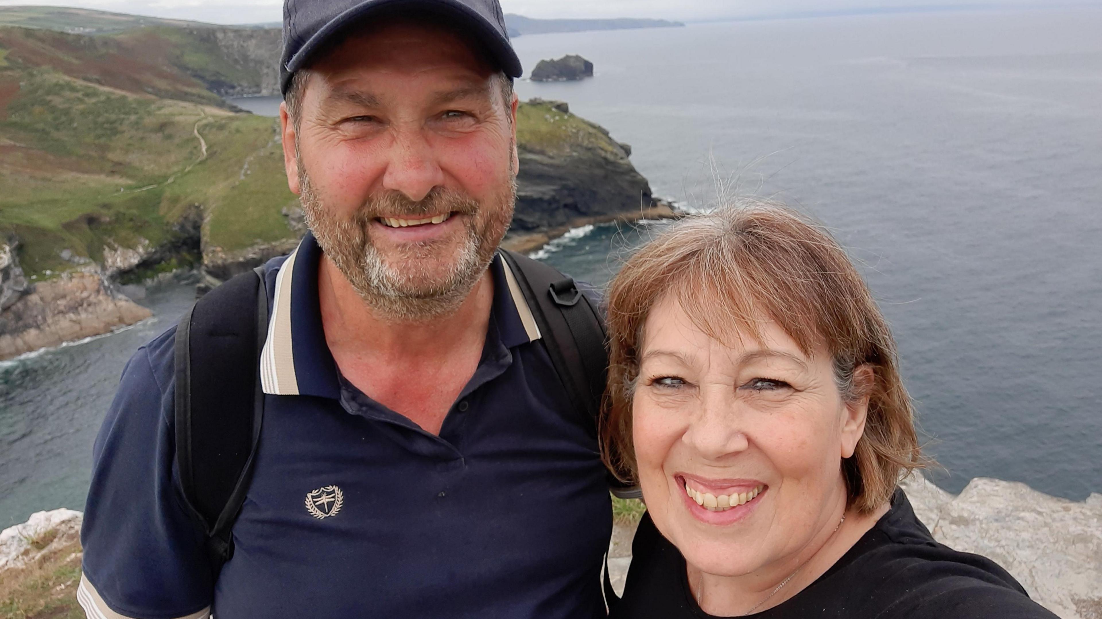 Kim and Lynda Sawyer smile for a selfie as they stand on cliffs with a view of the sea and coastline behind them. Kim, who has a beard, is wearing a dark blue polo shirt, a dark blue baseball cap and a black backpack. Kim, whose hair is cut in a bob, is wearing a black top.