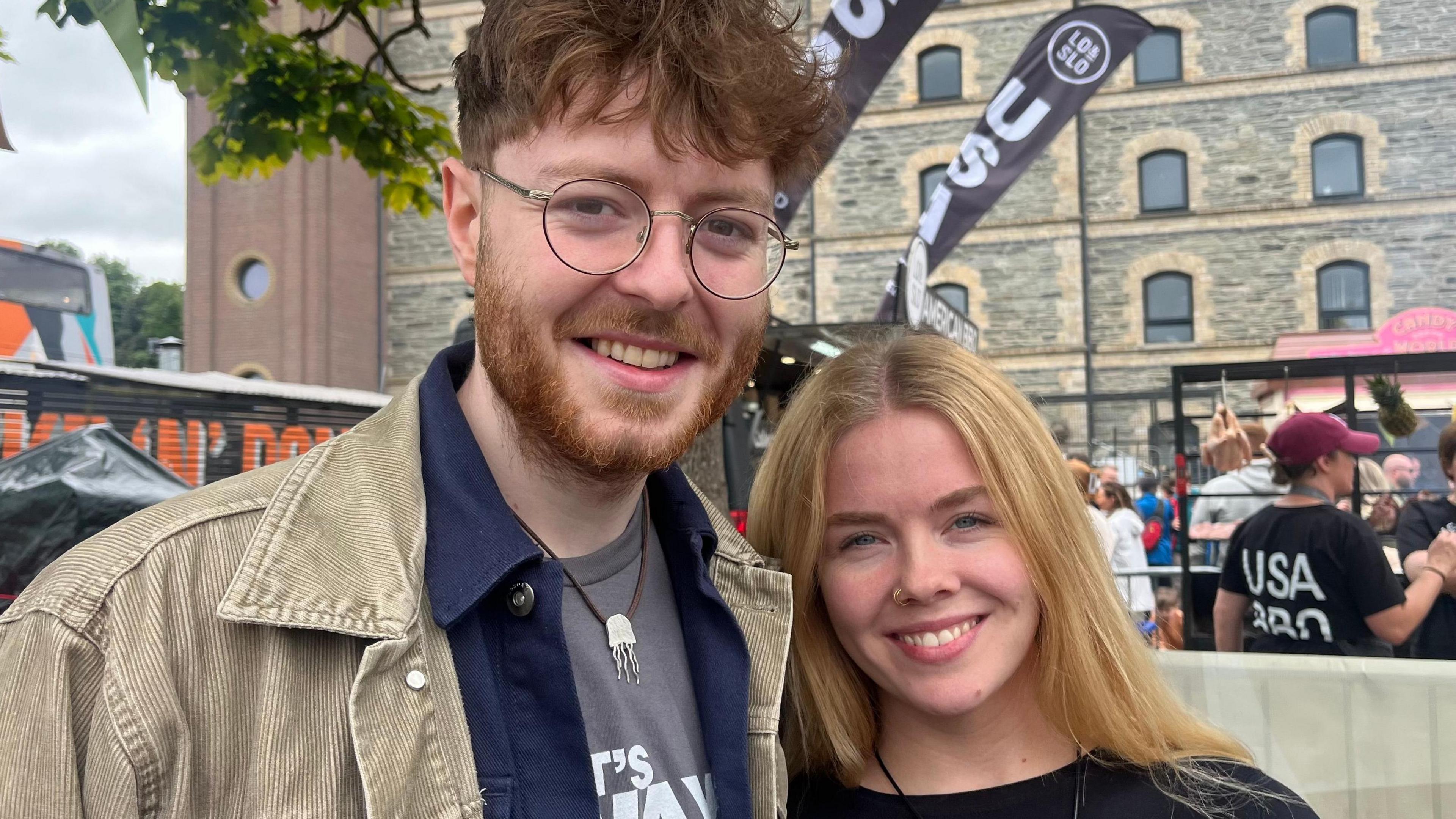 A man and woman standing close to each other in the centre of the photo. The man has brown hair and glasses and the woman has blonde hair. 