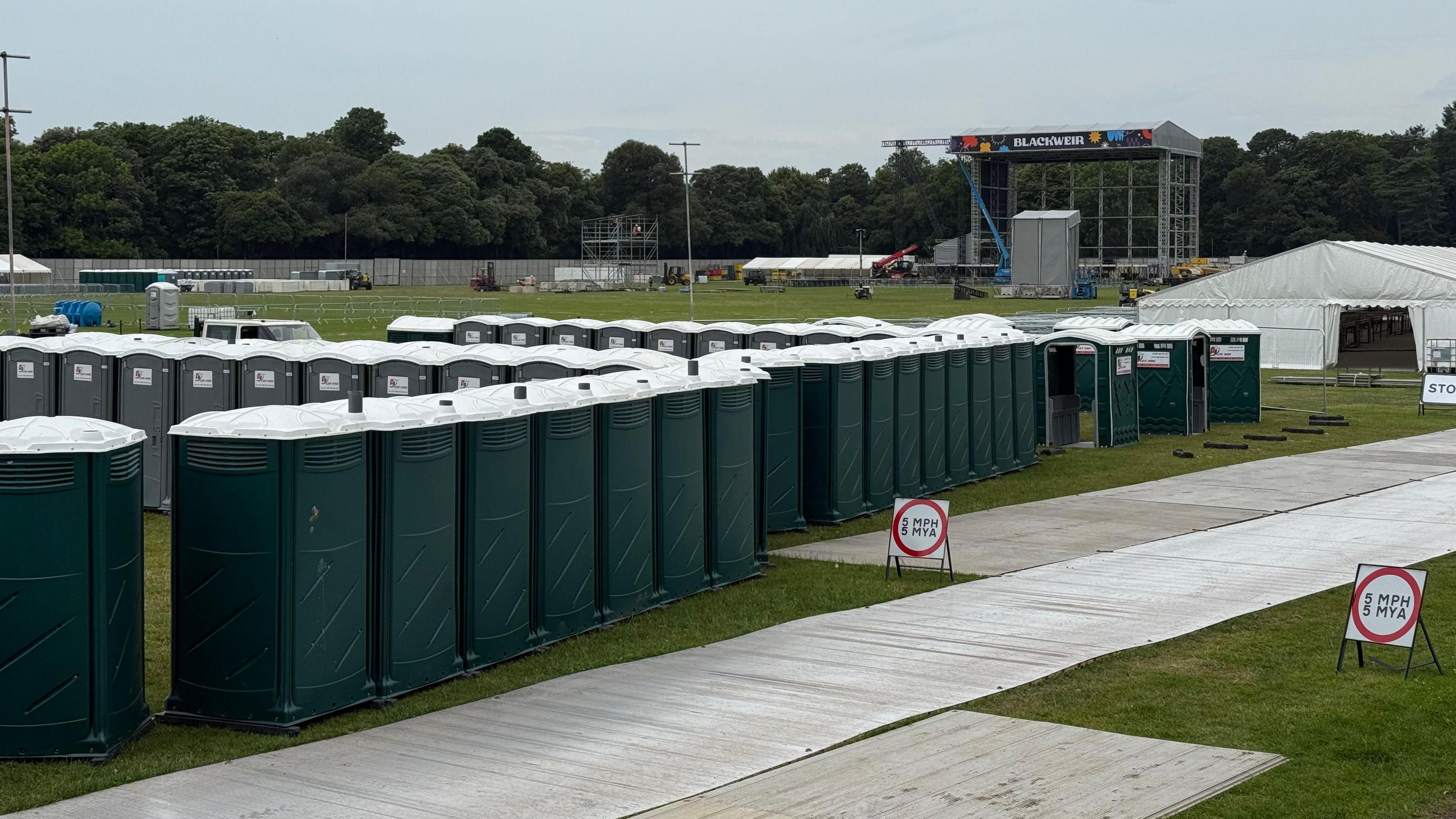 A row of portaloos can be seen in front of a stage being set up with the Blackweir name seen at the background attached to a state. There is also a white tent on the right of the photo. The are plastic walkways on the grass.