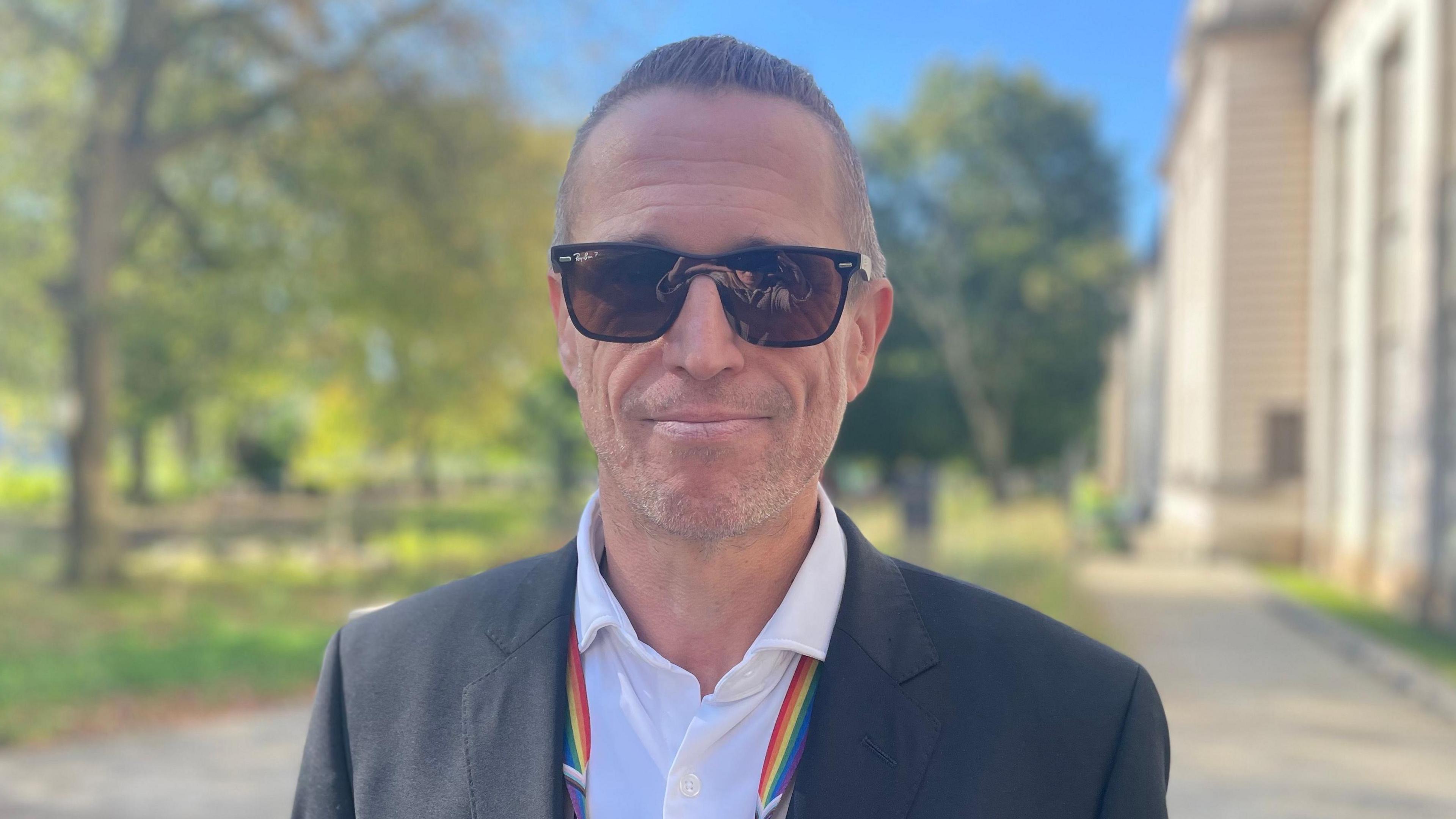 A man wearing sunglasses in a white shirt and dark grey jacket with trees and a university building in the background