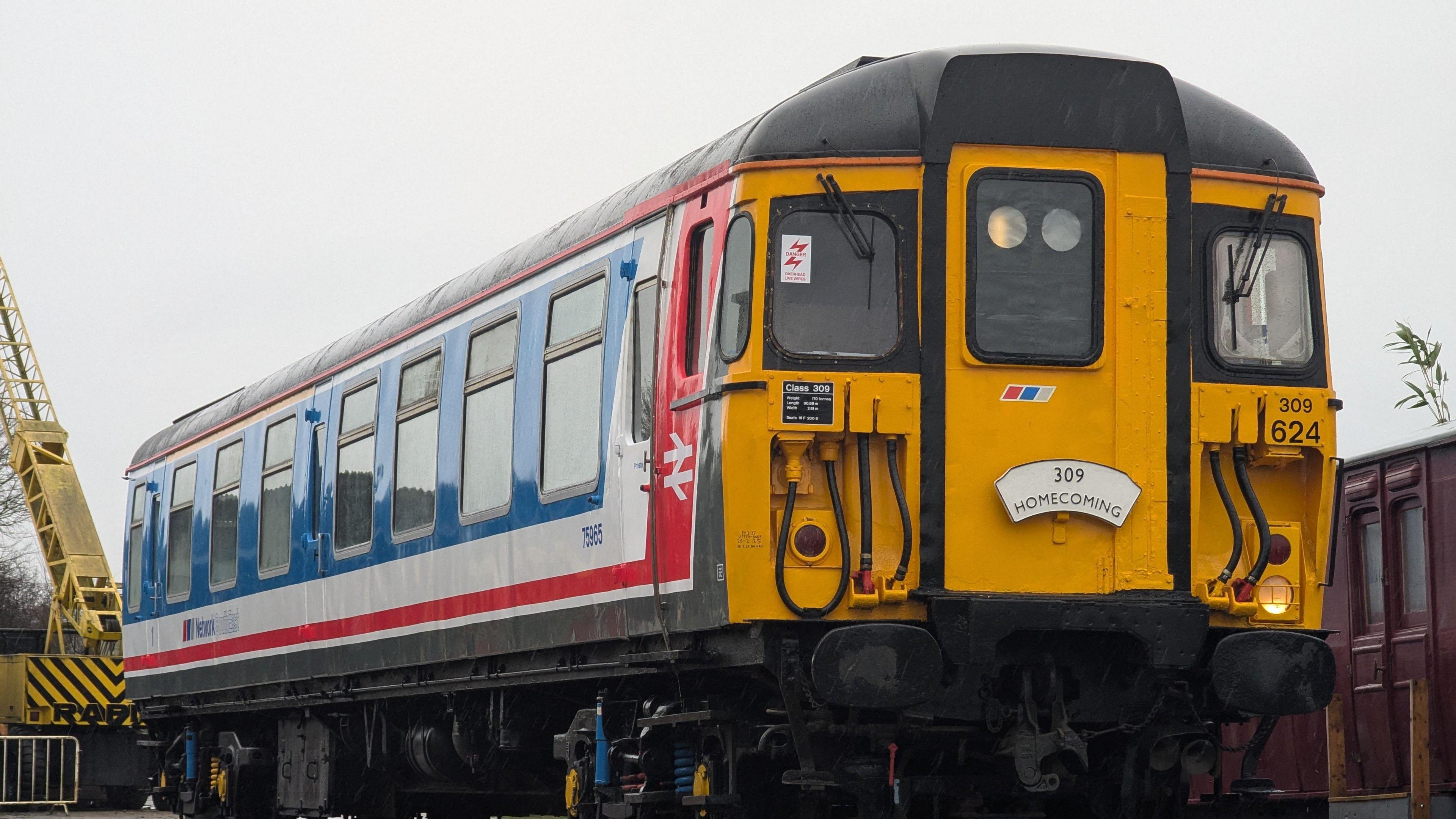 The Clacton Express pictured after the restoration works on its exterior. The electric train has a yellow front and a sign that reads: "309 Homecoming." On its side it is painted blue with white and red detailing. A crane can be seen behind the vehicle.