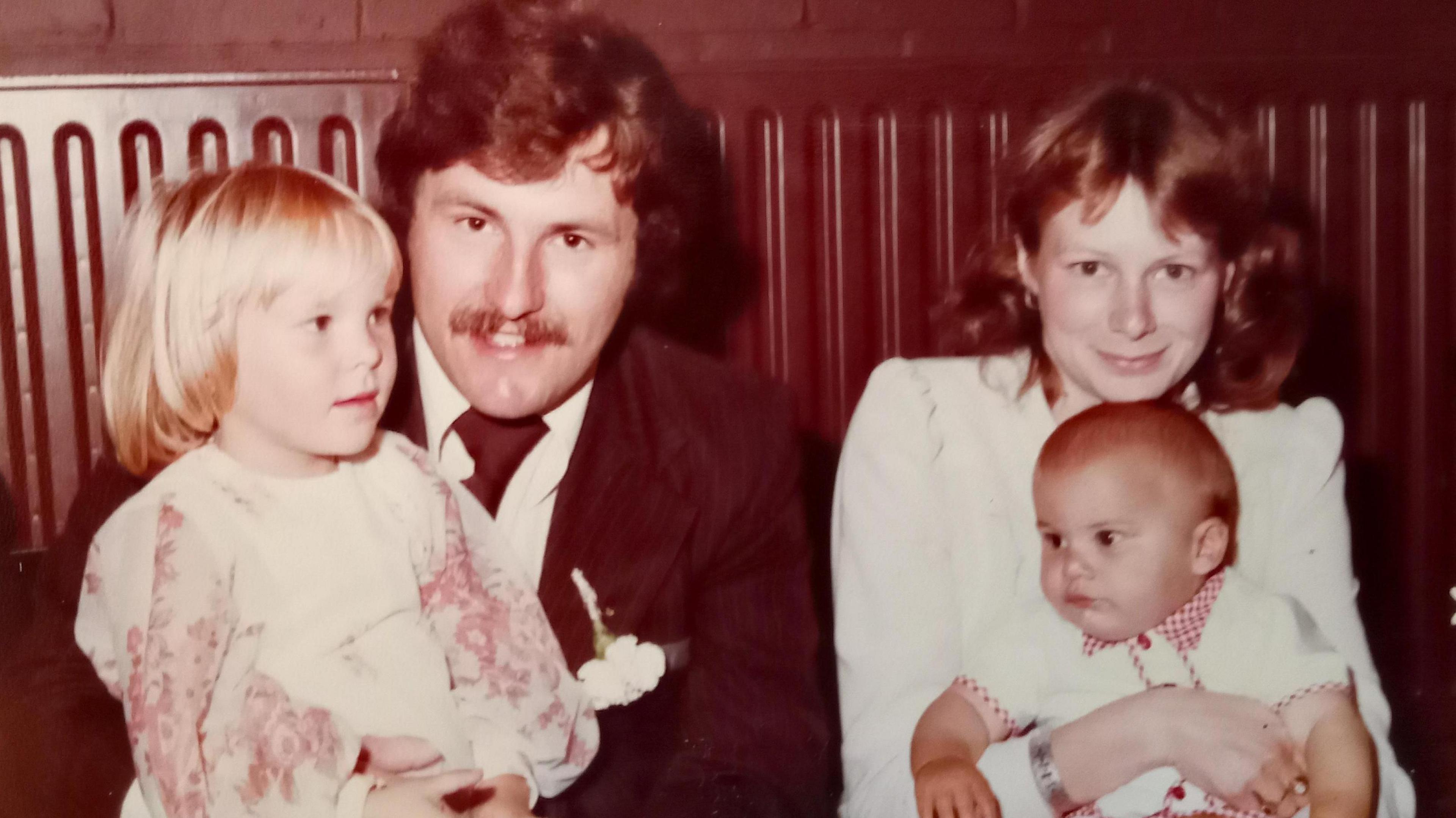 Black and White photo of Ray with toddler daughter Emma on his lap, sitting next to Anne who is holding their baby son Daniel. 