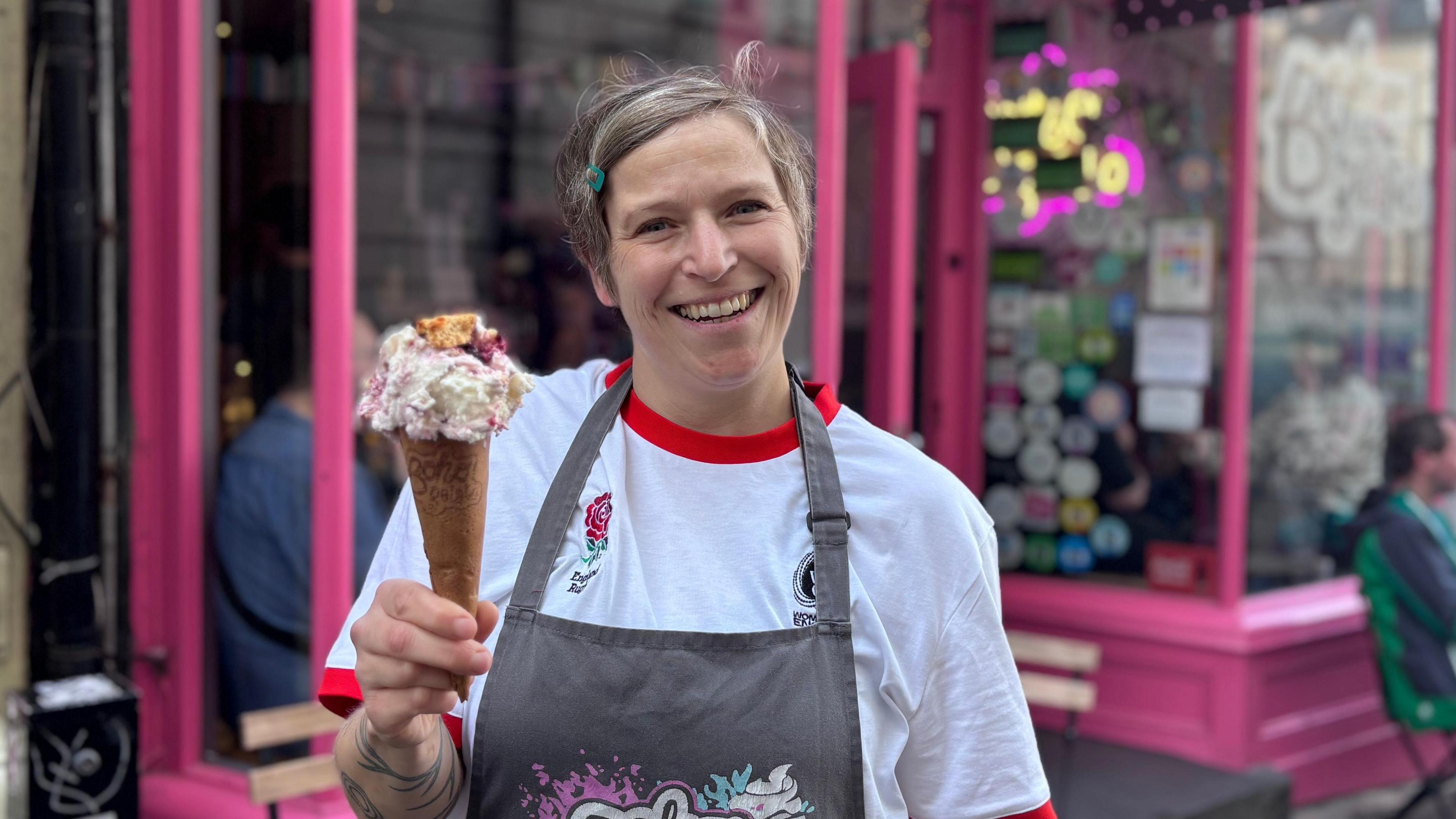 A women with short hair smiling at the camera. She is wearing a red and white England rugby T-shirt and is holding an ice cream cone with white ice cream on top. She is standing in front of a pink shop front.