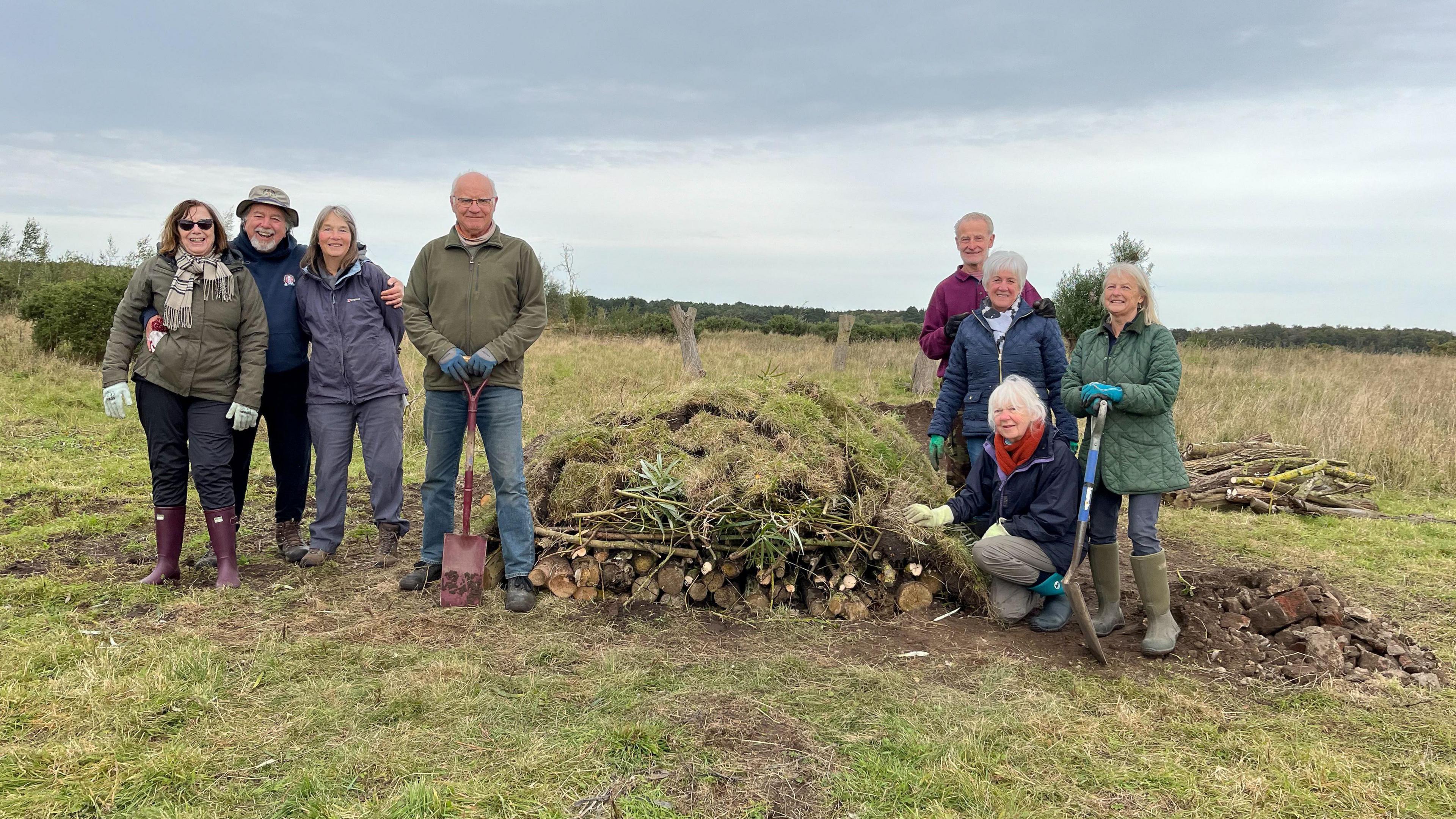 Volunteers of Lincolnshire Wildlife Trust at the nature reserve. A group of eight people standing together in a grassy field. The sky is cloudy. They are wearing outdoor clothing, gloves and boots and are smiling at the camera. In front of them is a habitat pile made of logs, branches and turf. 