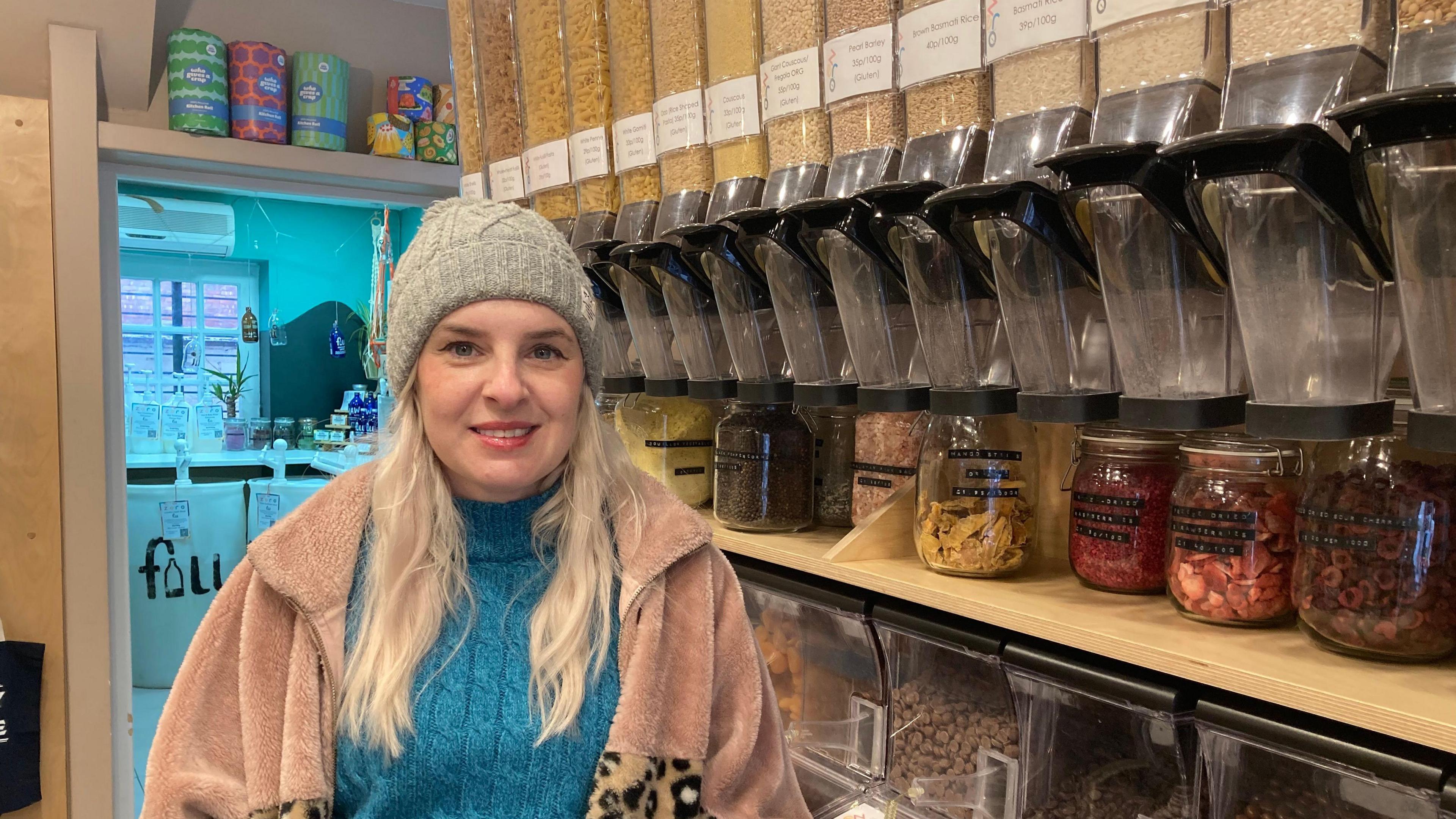 Owner Charlie Demetriou is wearing a woolly hat and a fleece jacket as she stands in front of stock in the store