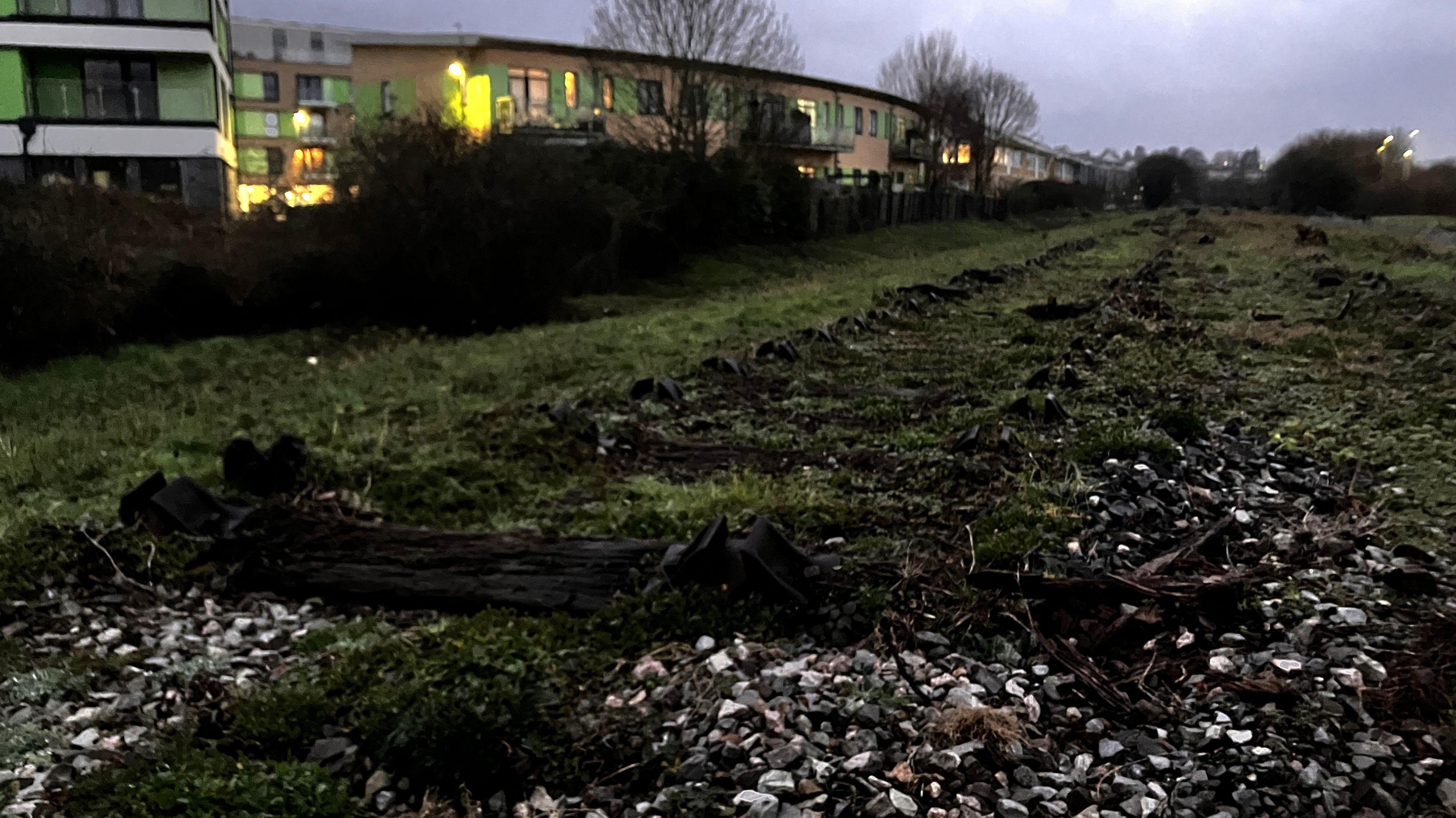 An old railway line with rocks and grass growing over it and a large building in the background