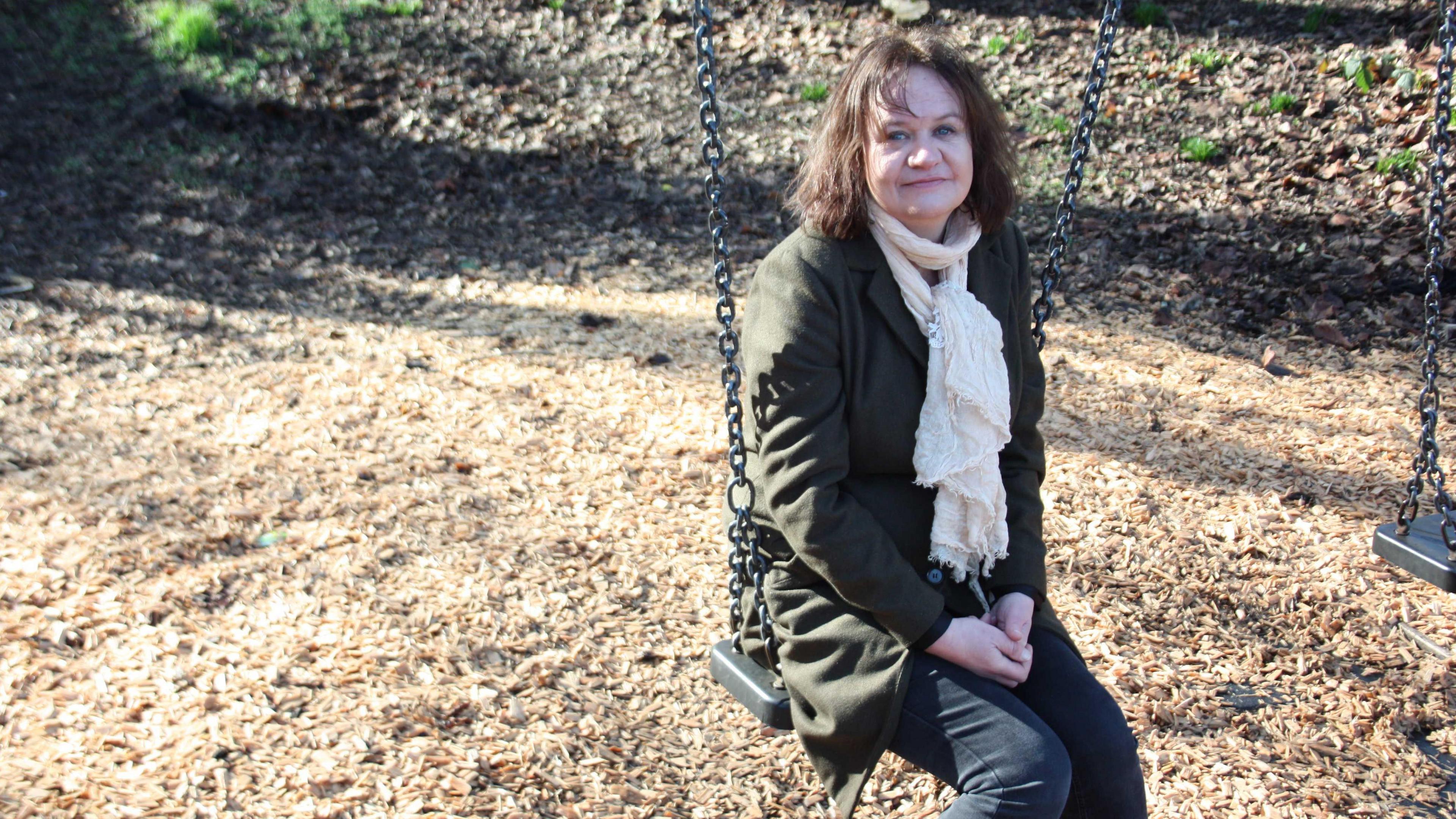 Dr Sara MacRae sitting on a swing with light brown bark covering the ground unerneath and a bank of earth in the background. She is wearing a green coat and a cream scarf.