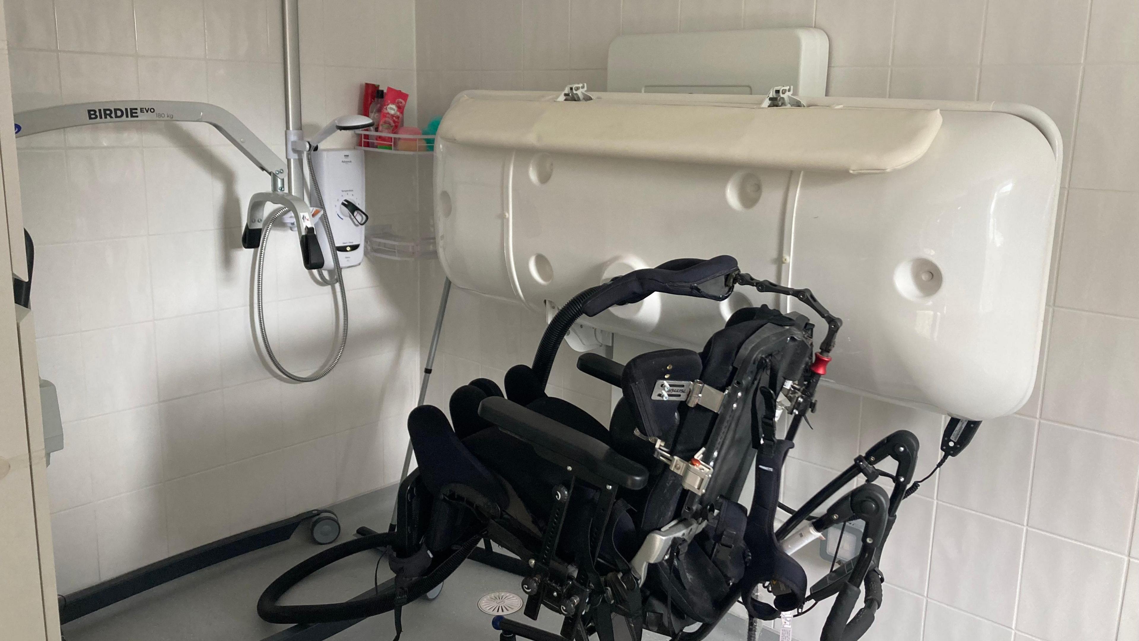 A wet room with shower, hoist and changing table. The tiles are white and the floor grey.