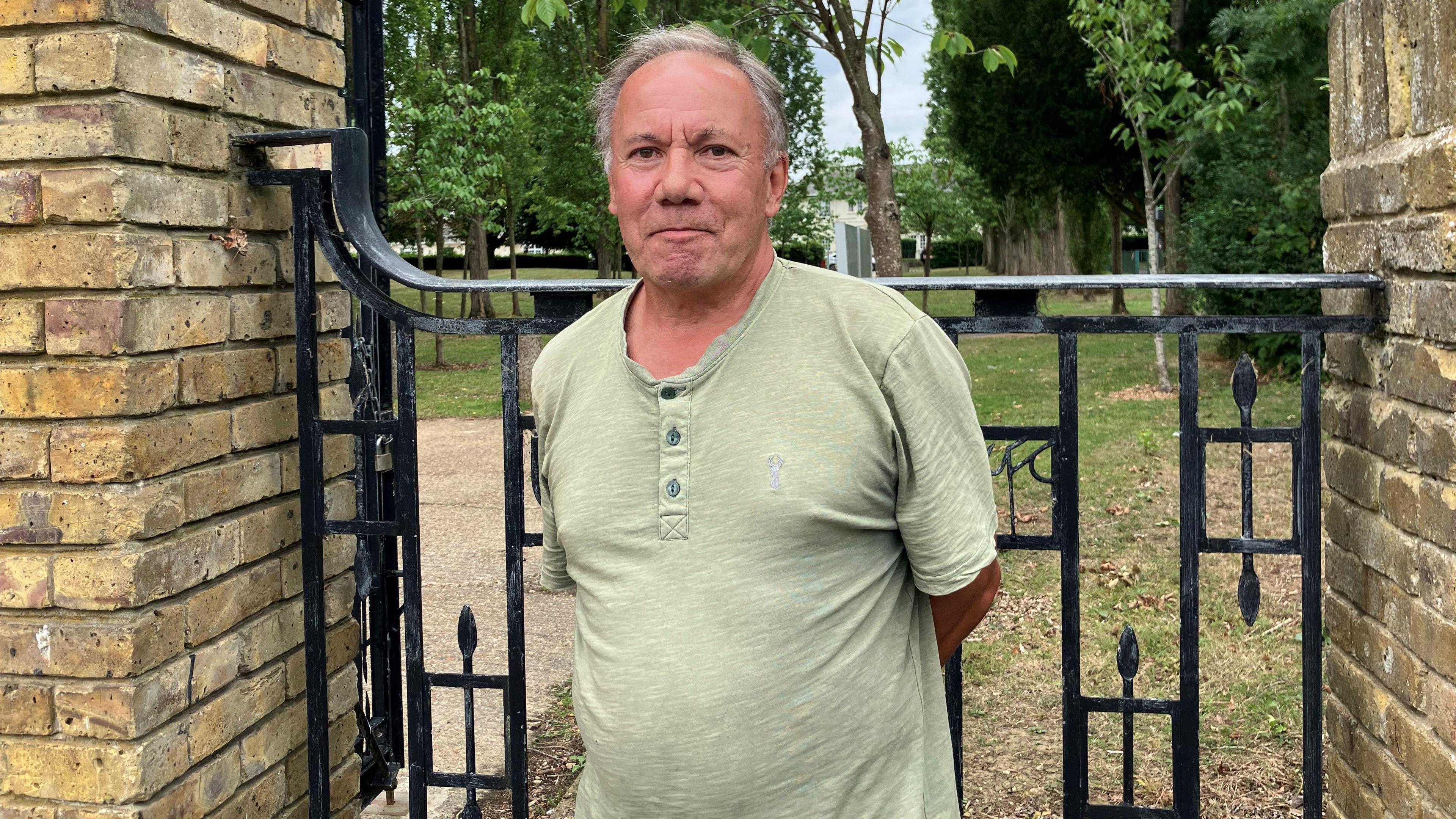 Paul Hayden looks directly at the camera and smiles. He is wearing a green T-shirt and is standing in front of a black gate with a patch of grass behind it.