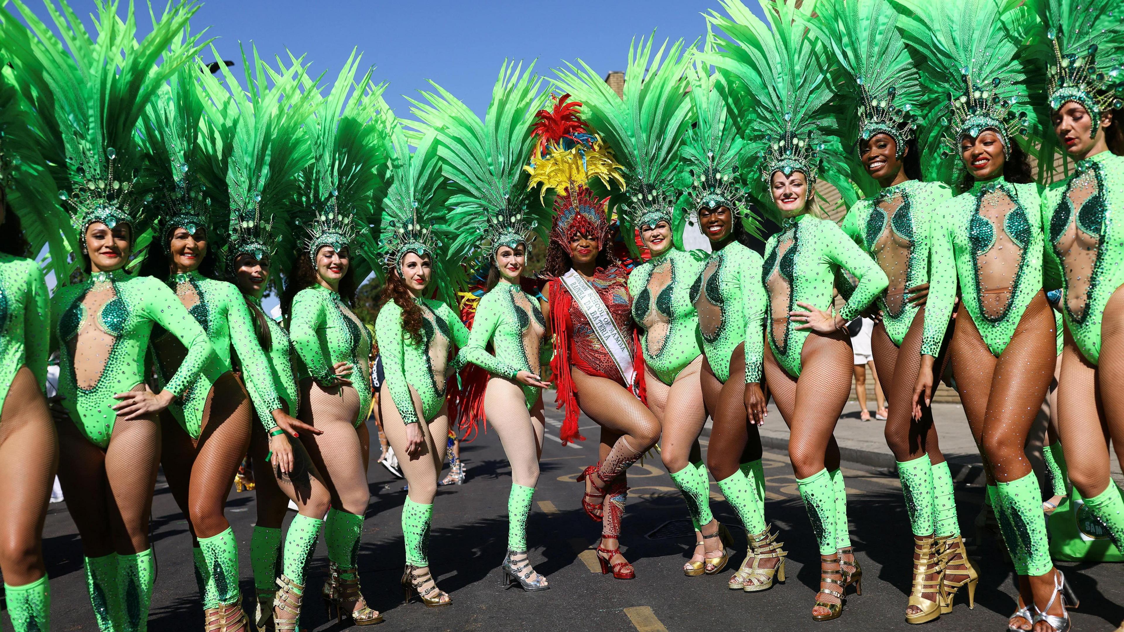 A group of women stand in a row in green leotard costumes, with a woman in red in the middle. Their outfits feature sequins, gems and feathers.