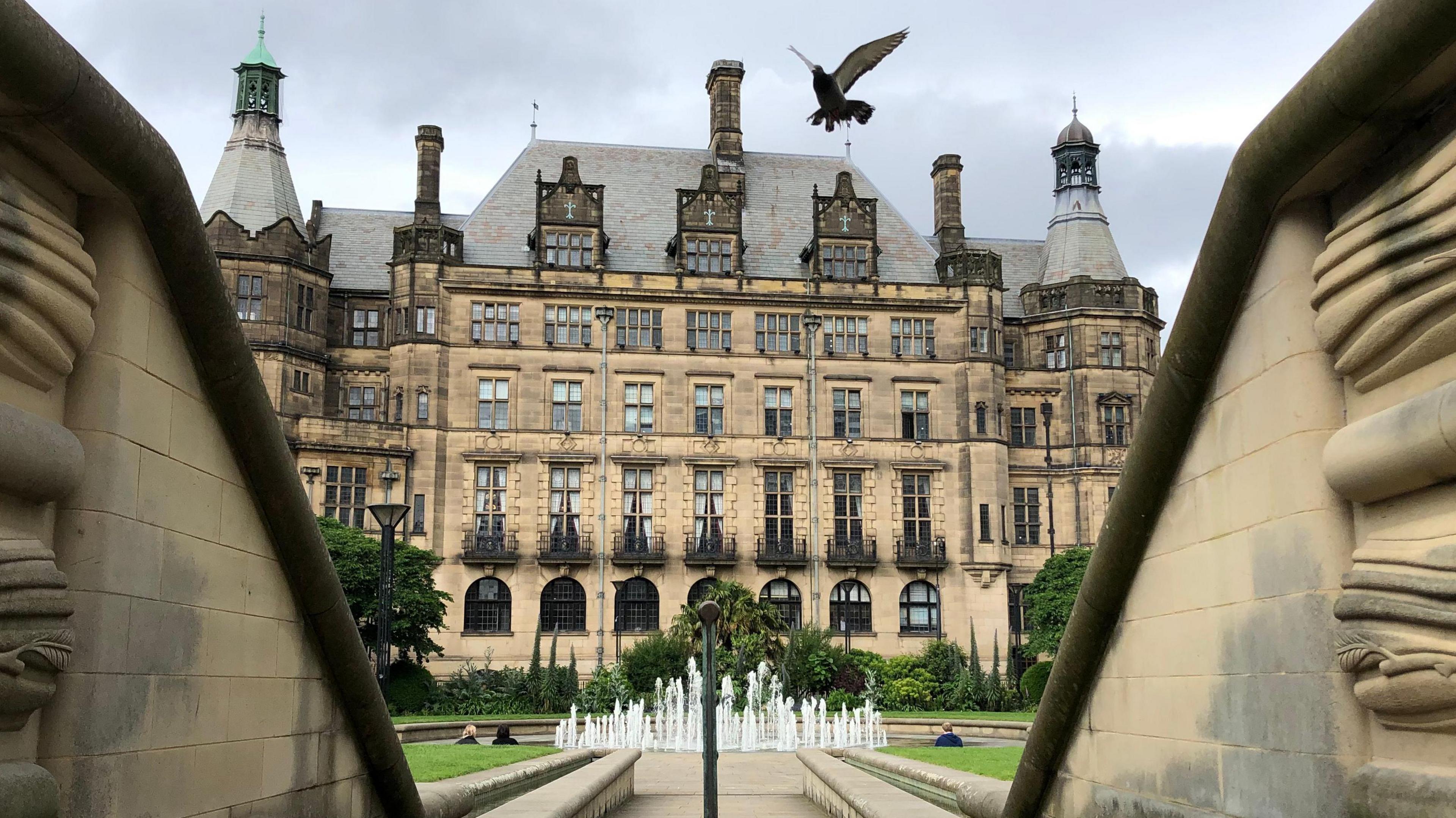 A grand five-storey gothic building with turrets at each end. In front of the building is a fountain and few bushes. Three people are sat around the fountain. In the foreground, the view of the building is framed by two stone low walls.