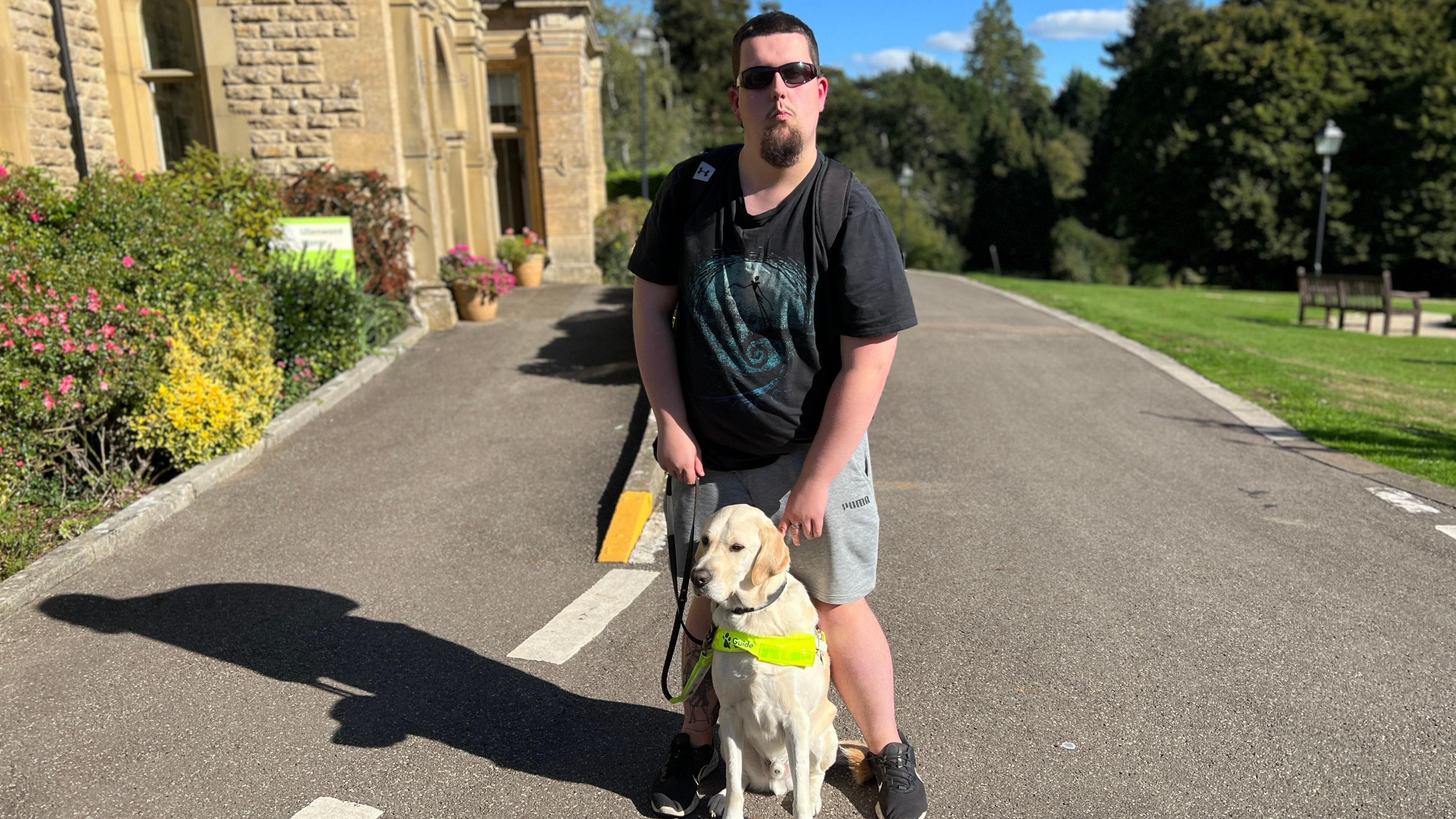 Chris is wearing dark glasses and a black t-shirt with shorts. He has a yellow Labrador service dog between his legs. Chris has dark hair and a small beard.