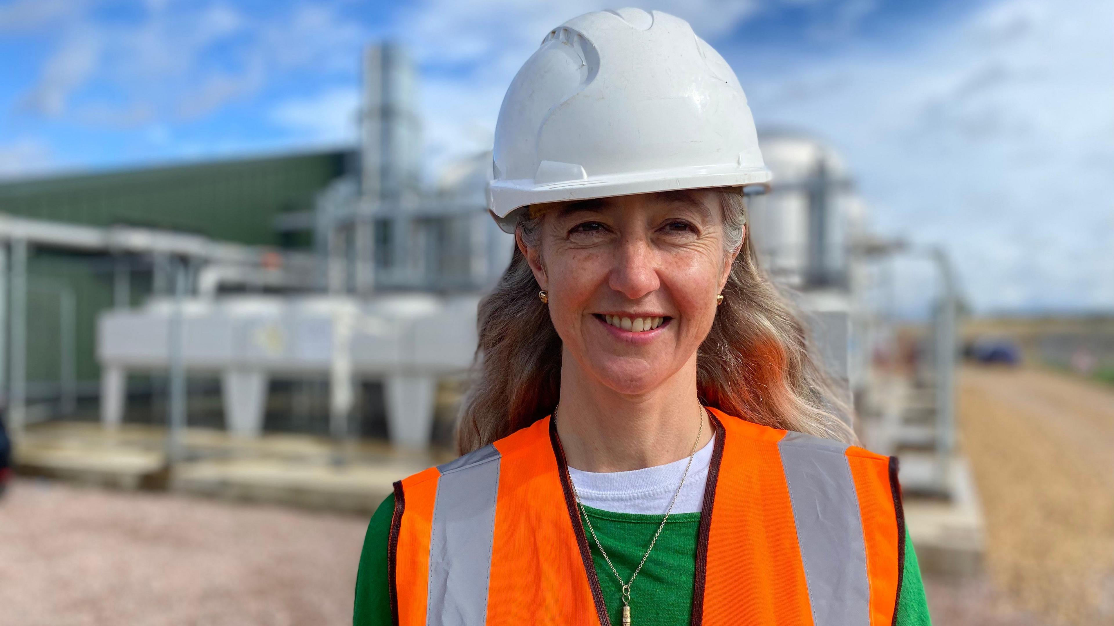 Headshot of Ros Hathorn standing in front of the heat network infrastructure, which is a set of pipes and tankers behind wire gates. Hathorn is wearing a white hard hat and orange hi-vis vest. She has long light hair, is smiling and wearing a small necklace with a green jumper and white top underneath.