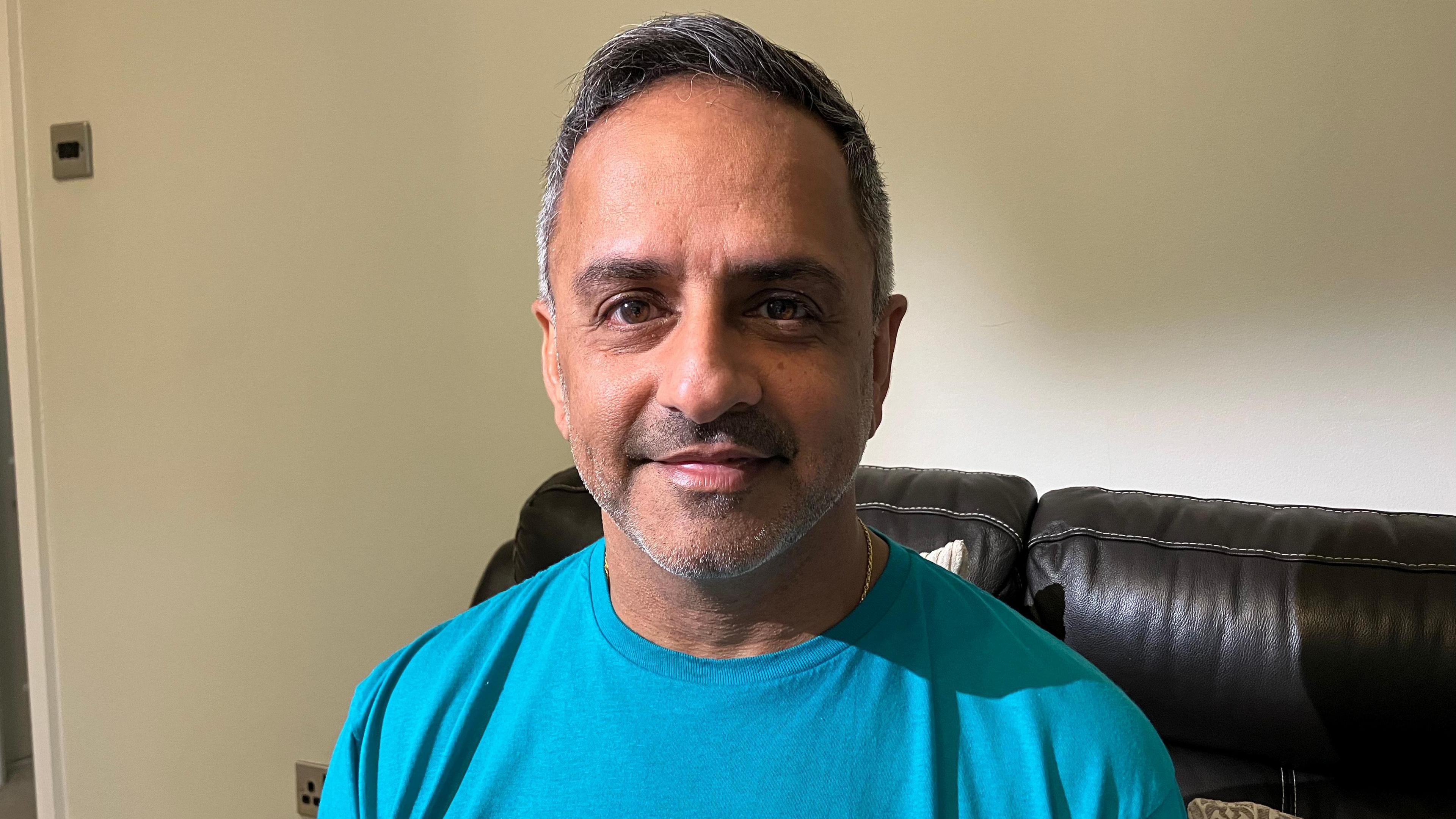 A man in a blue t-shirt sitting on a brown leather sofa. A cream wall can be seen behind.