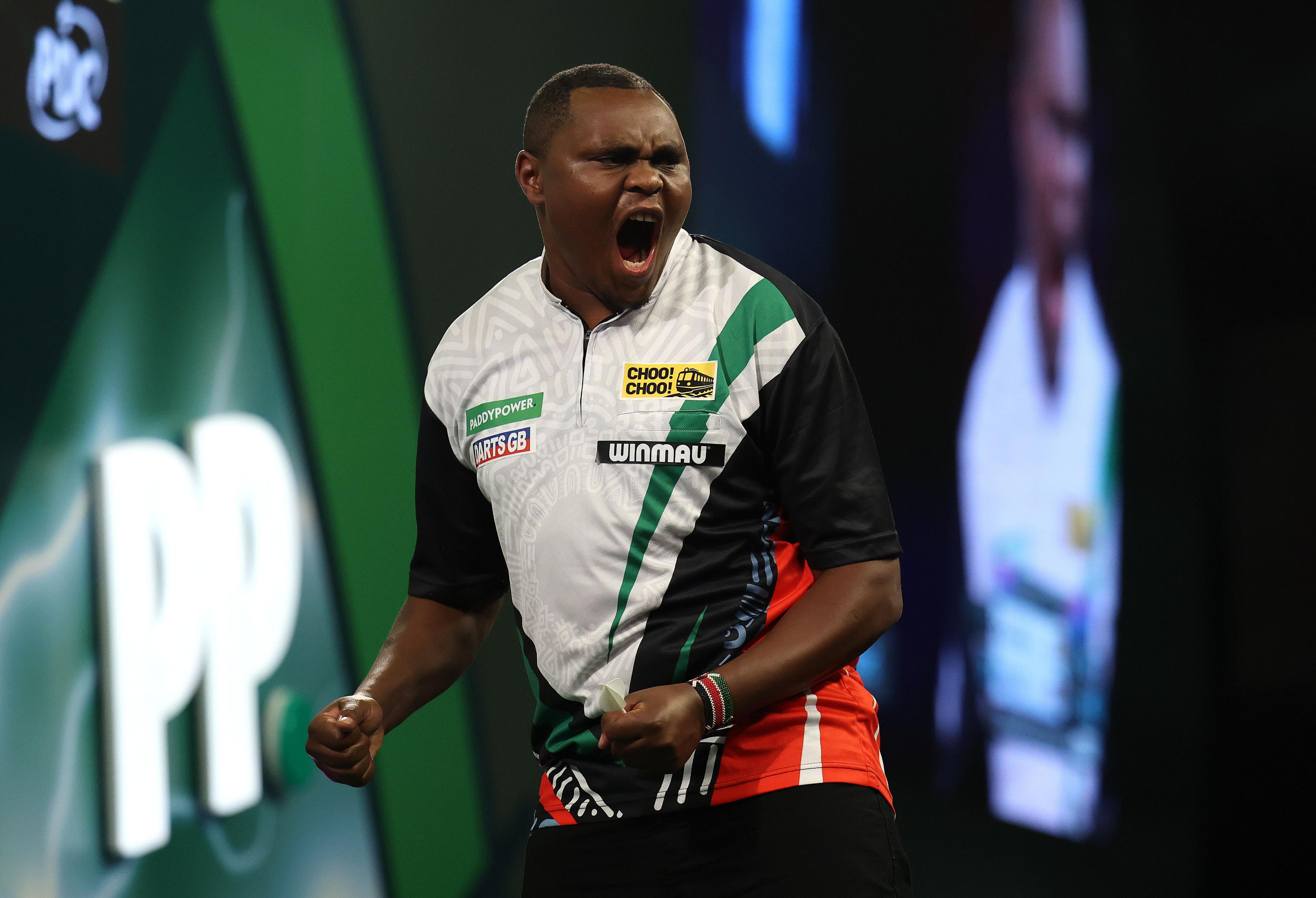 A darts player wearing a multicolored shirt featuring green, white, black, and red panels stands on stage with a clenched fist, appearing to celebrate. The shirt displays several logos, including “CHO CHOO!” and “WINMAU.” The background shows a large illuminated “PP” sign and a blurred screen projection of the player.