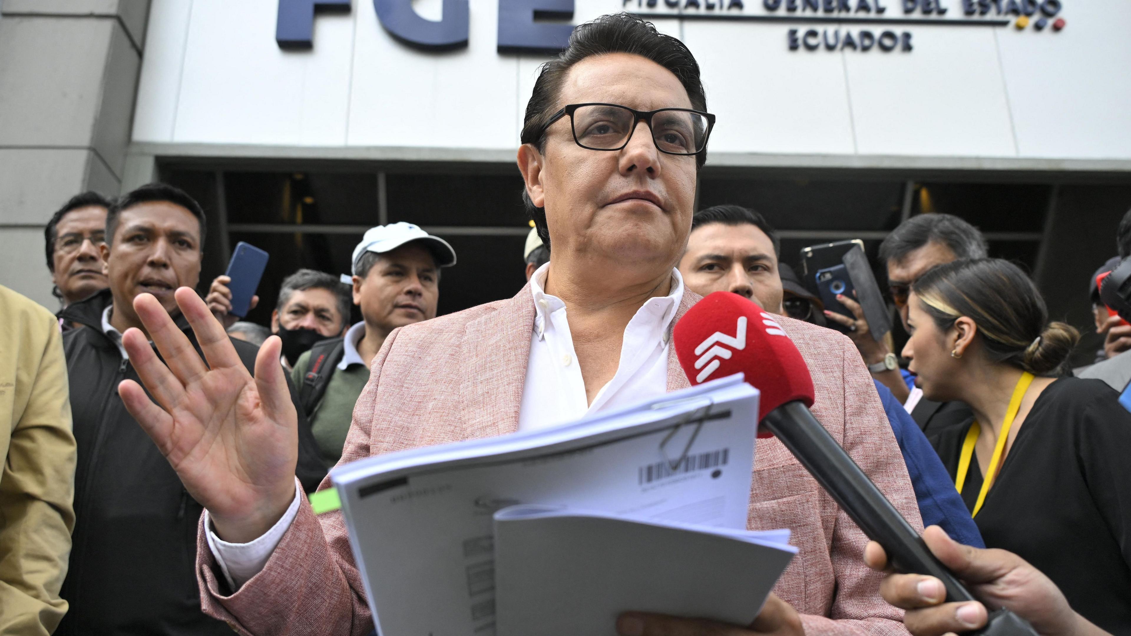 Fernando Villavicencio speaks into a microphone while holding papers and gesturing with one hand on the steps of a building surrounded by people. He wears a pink jacket, white shirt and lack-rimmed glasses.