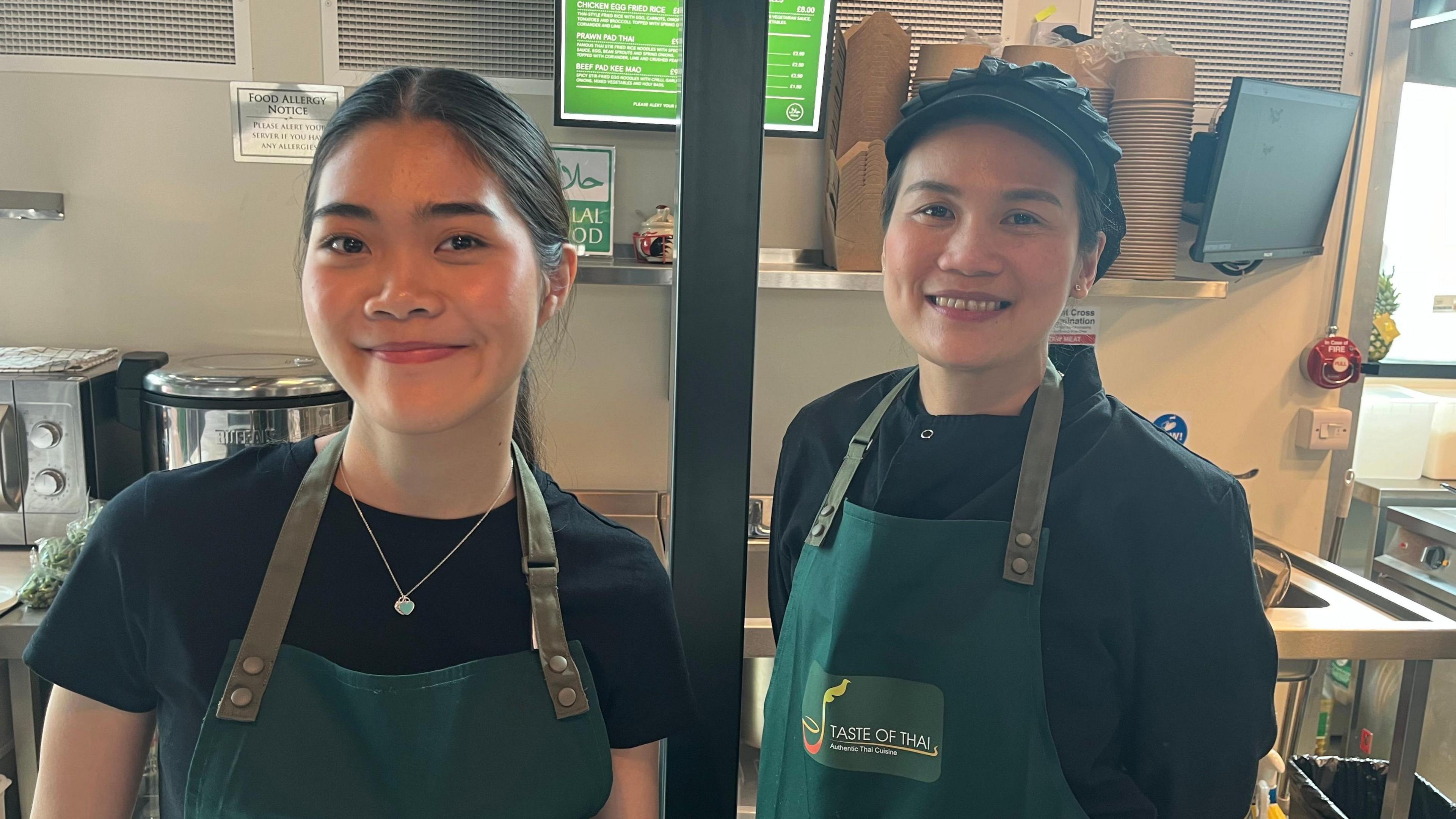 Two women smiling wearing aprons with kitchen behind them.