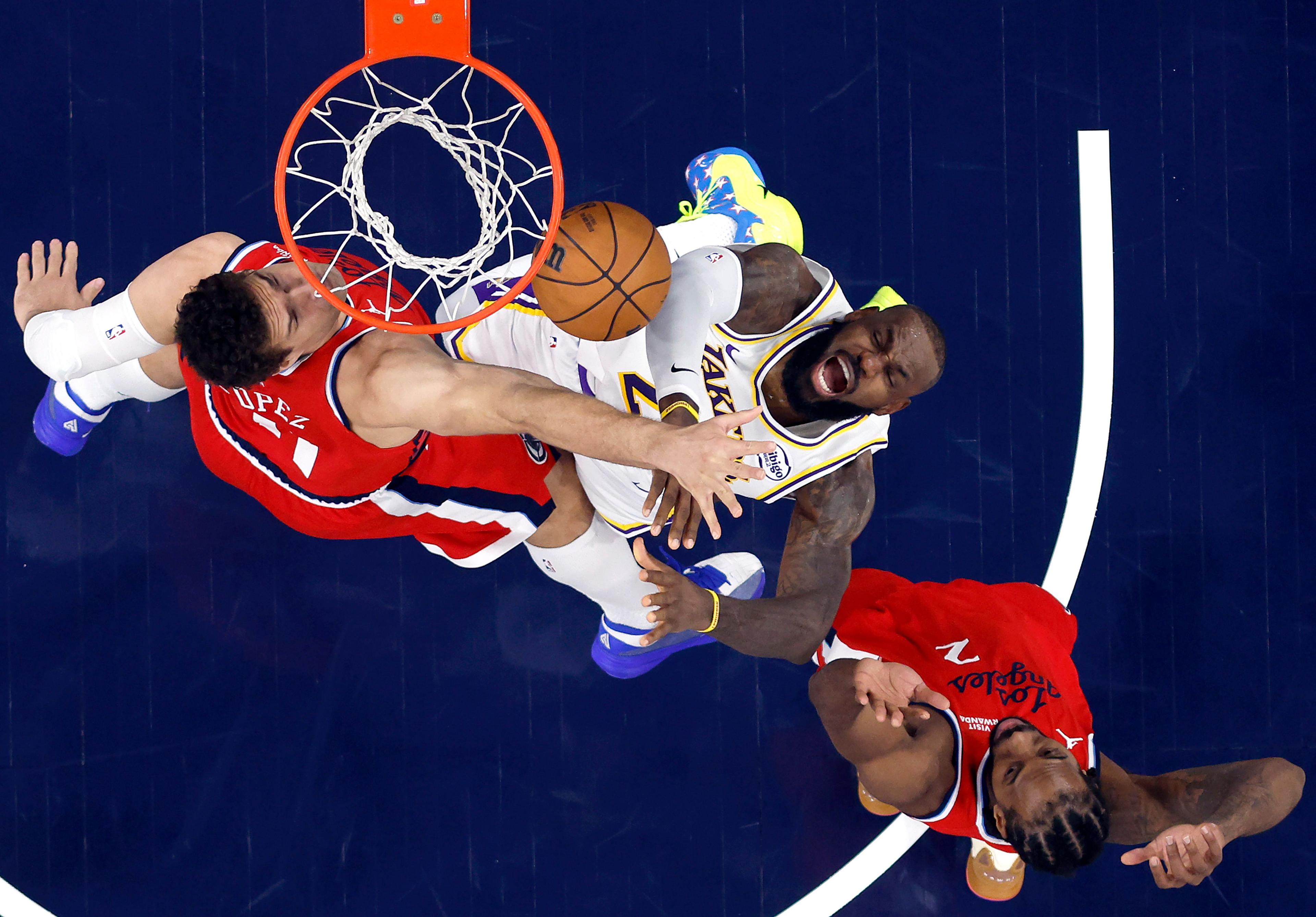 An overhead view of a basketball game shows three players near the hoop. One player in a white and gold uniform is attempting a shot while being closely defended by a player in a red uniform. Another player in red is positioned nearby, looking toward the action. The basketball is just above the rim, and the dark blue court contrasts with the players’ bright uniforms.