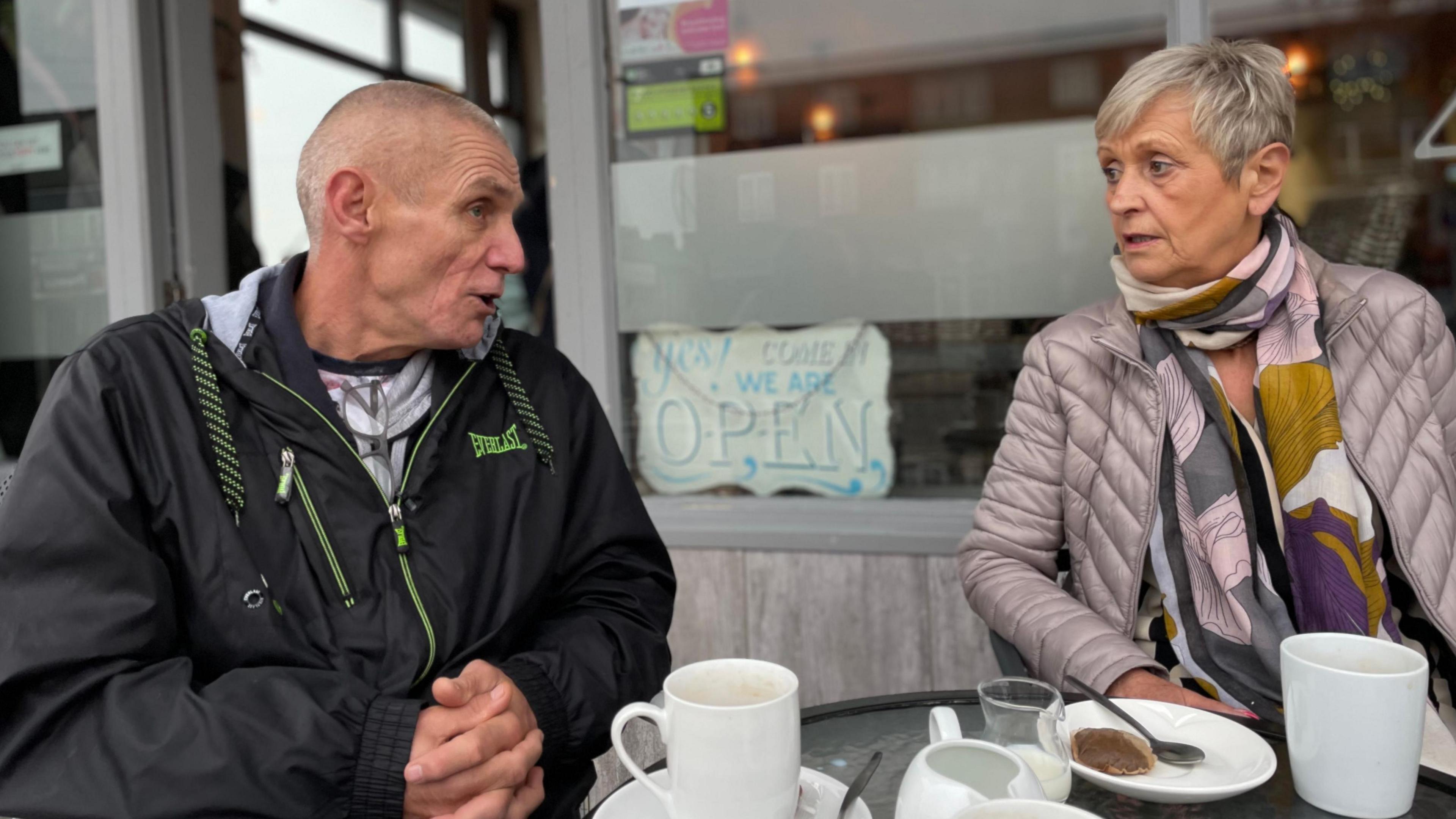 John Taylor and Barbara Thornton talk as they sit at a table outside a cafe in Scunthorpe. Mr Taylor has close-cropped grey hair and wears a black rain jacket. Mrs Thornton has short grey hair and wears a silver-pink jacket and a colourful scarf. White porcelain cups and saucers stand on the table between them.