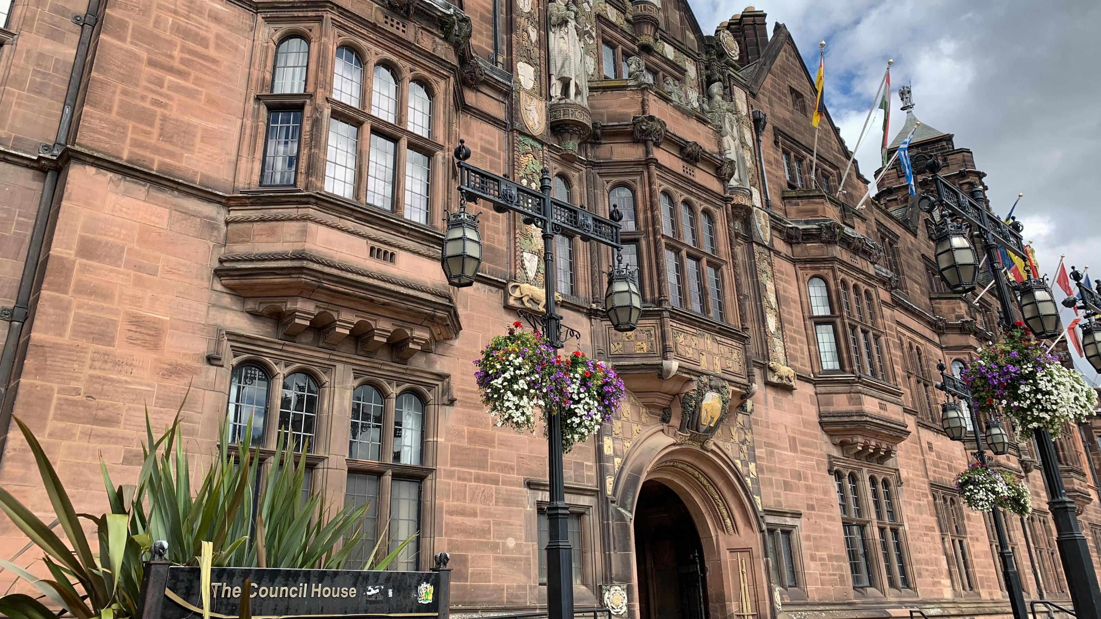 A picture of Coventry City Council's Council House. It's a Neo-Gothic building made of reddish sandstone. outside are colourful hanging baskets.