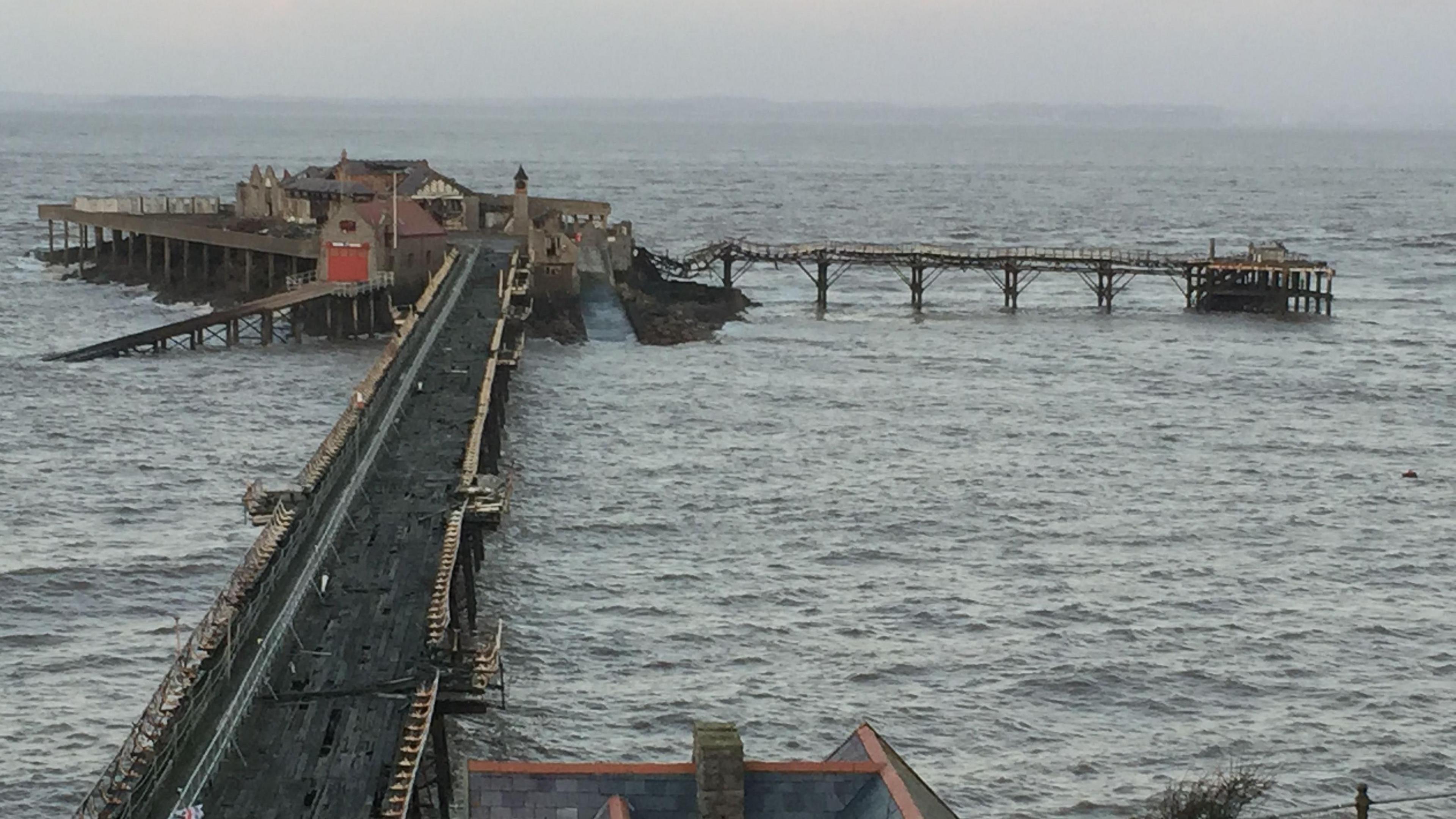 Tattered-looking pier in grey, rough seas. One of the walkways is twisted and warped, and the old red roller door of the previous RNLI station is visible, along with the ramp that takes the boats to the sea.