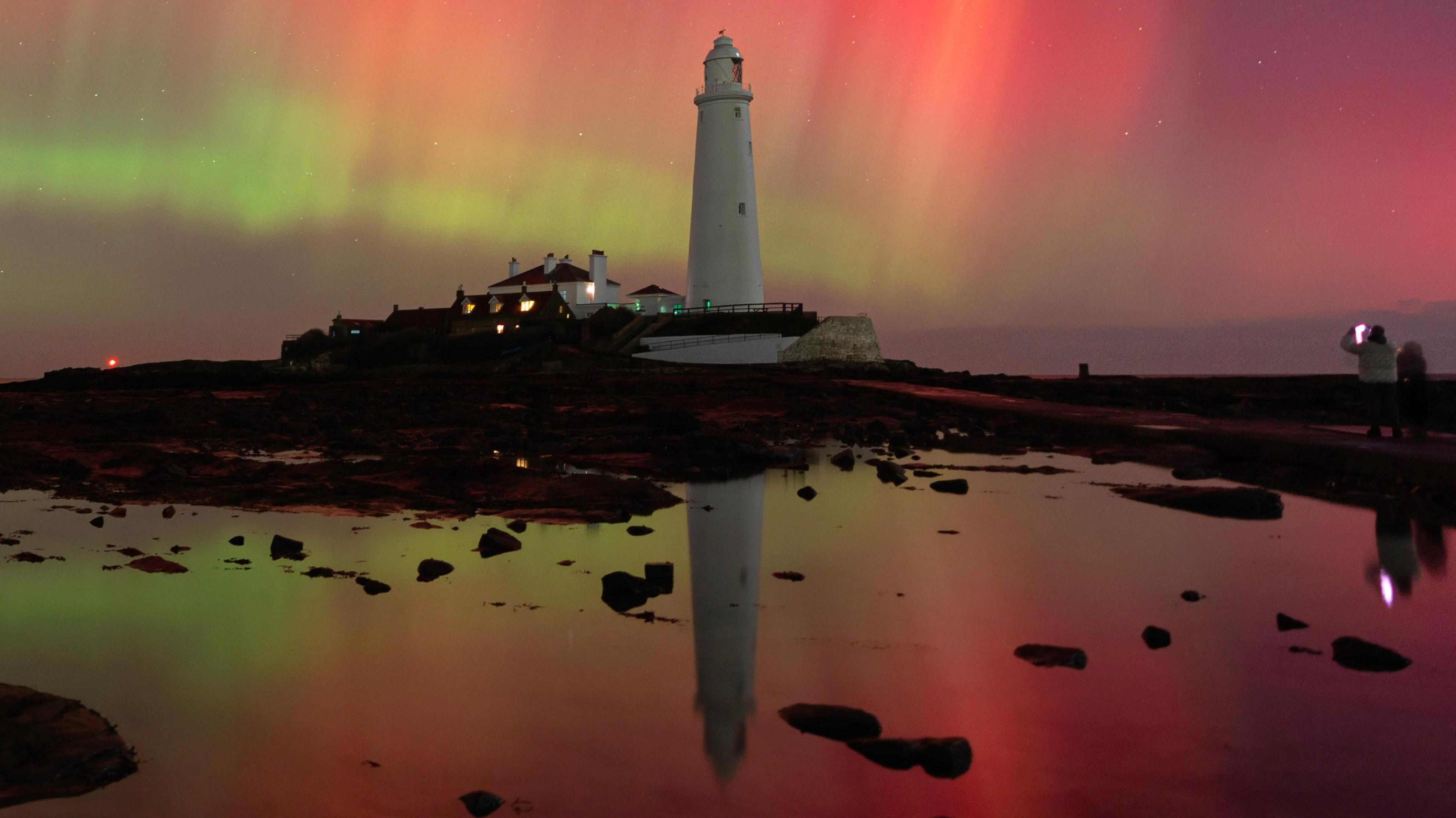A white lighthouse with a blaze of red and green lights in the sky behind it, all reflected in a pool of water in front