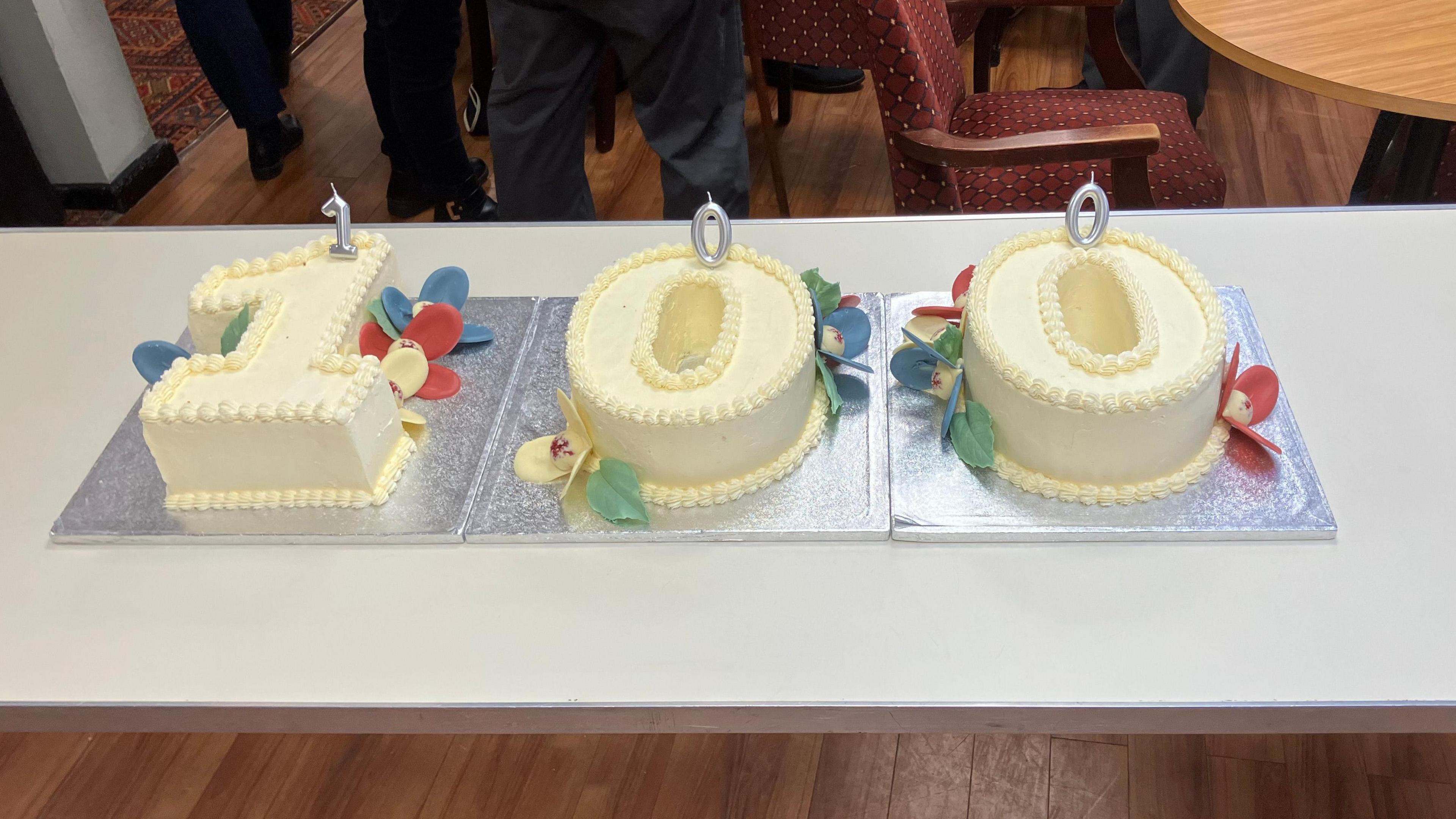 three iced cakes spelling out 1-0-0 -on silver platters on a table