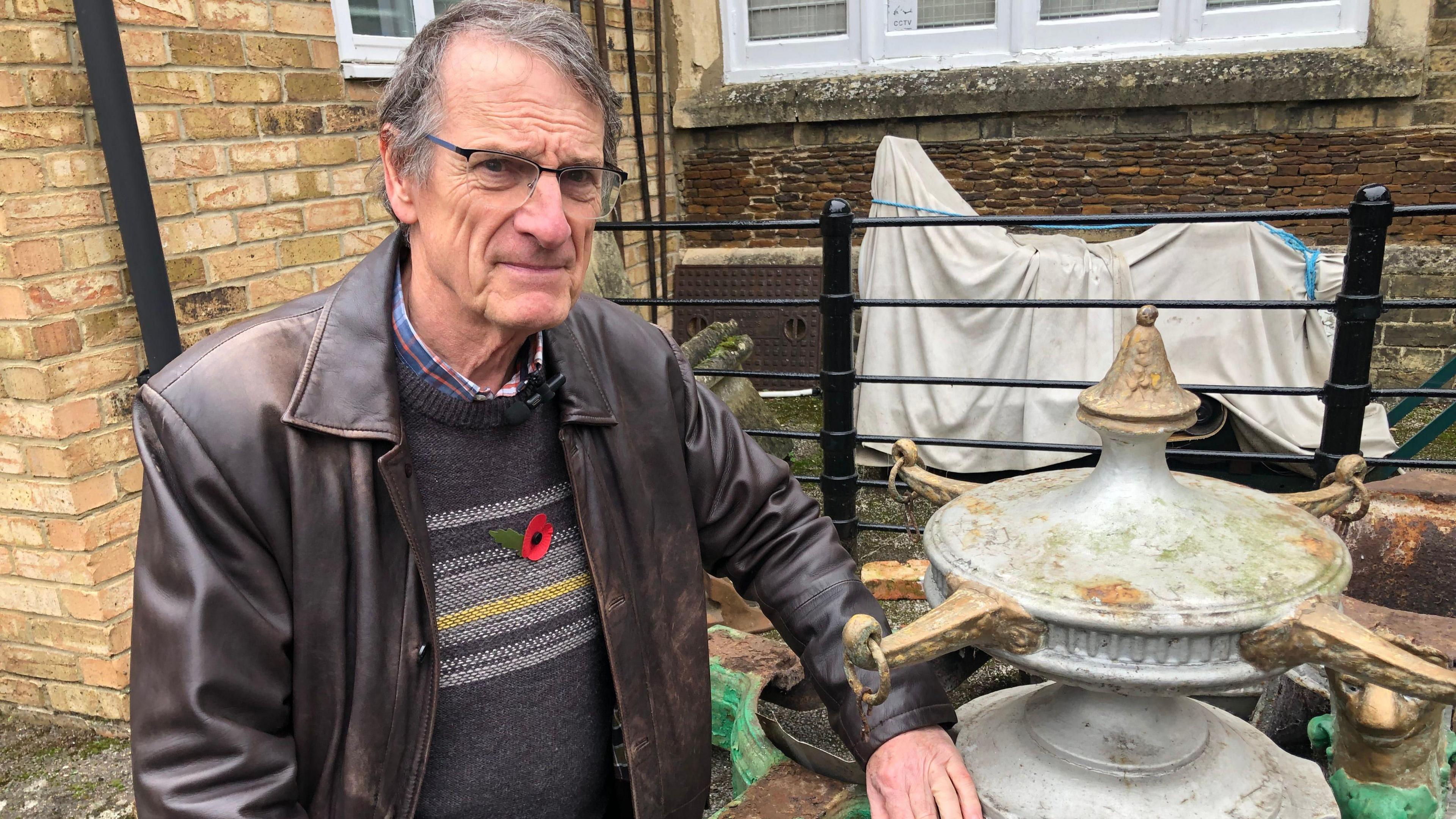 Gordon Thorpe is wearing a brown leather jacket and a dark jumper with a poppy in the middle. He has a chequered shirt underneath and is resting his left arm on part of the cast iron water feature, which is painted silver, with four thin gold arms sticking out around 15cm (6 inches).