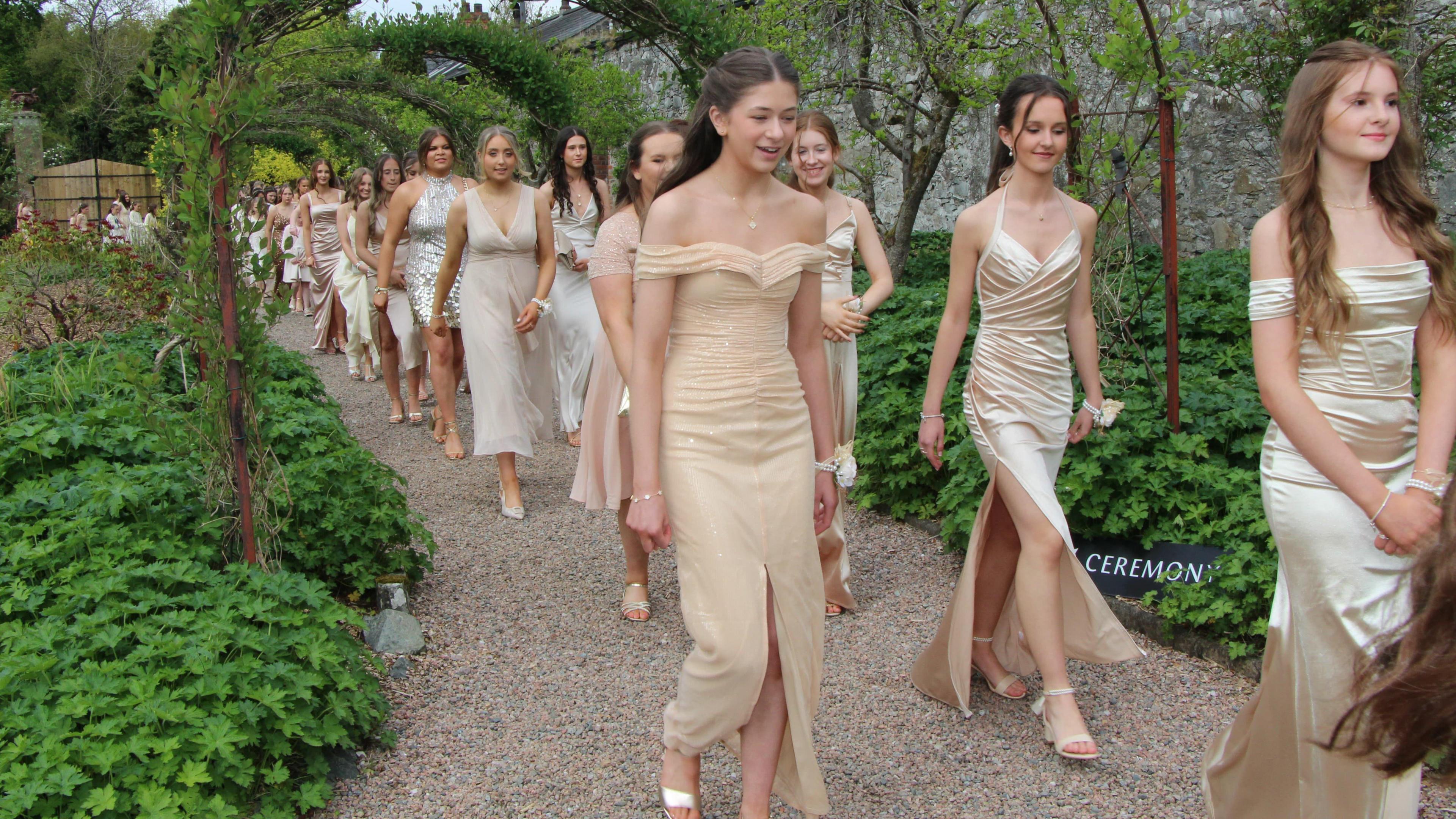 Bridesmaid walking down an aisle which has a garden arch above their heads. They are wearing gold dresses and walking through the arch.