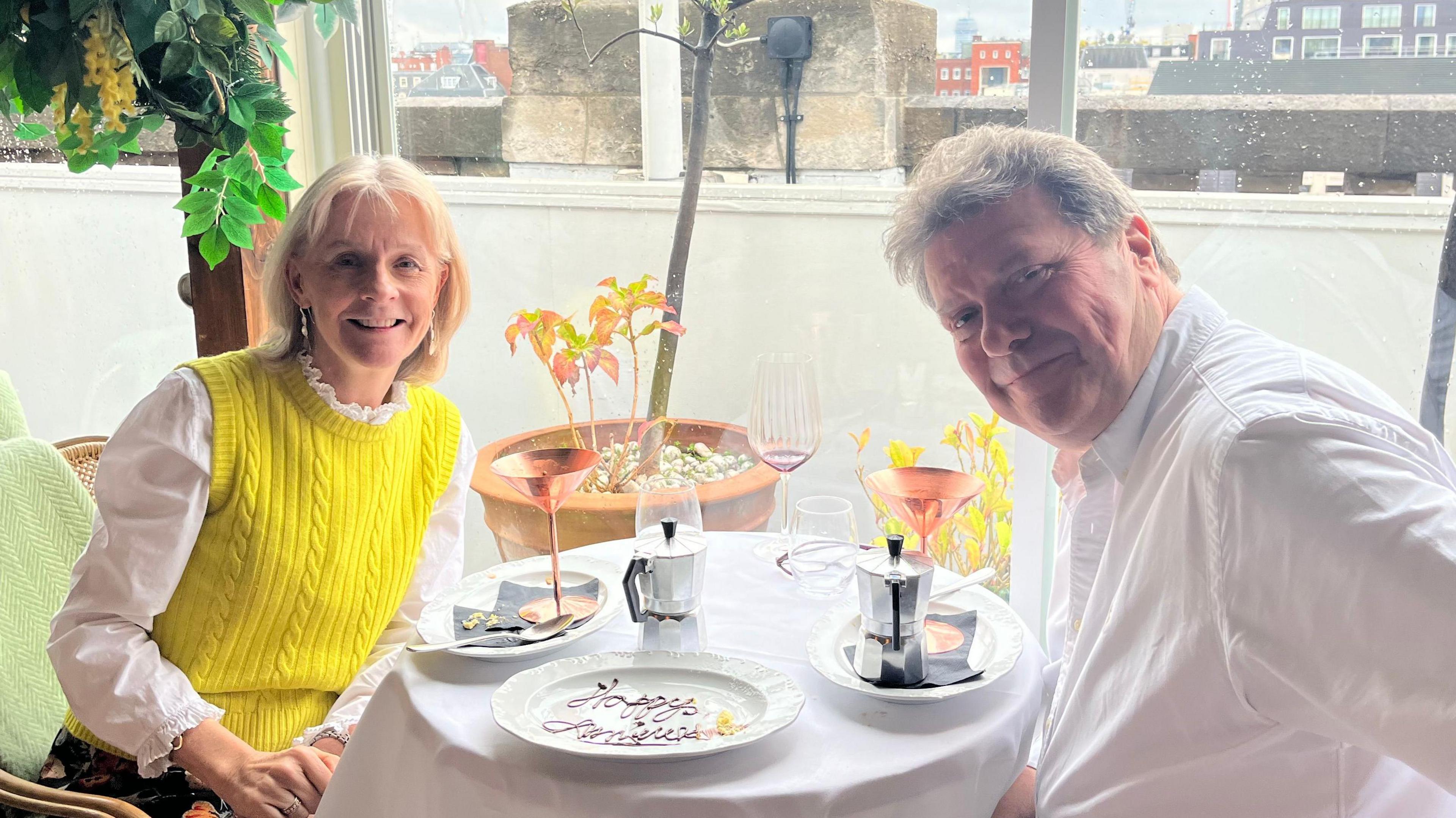 Sue and Alex sit at a table in front of a window, a plate saying 'Happy anniversary' in chocolate between them. They smile at the camera, Alex's arm outstretched Sue is wearing a yellow sleeveless jumper over a white shirt and earrings, she has blonde hair. Alex is wearing a white shirt and his hair as turned grey. There are two pots of what looks like coffee at the table and small plants and buildings in the background.