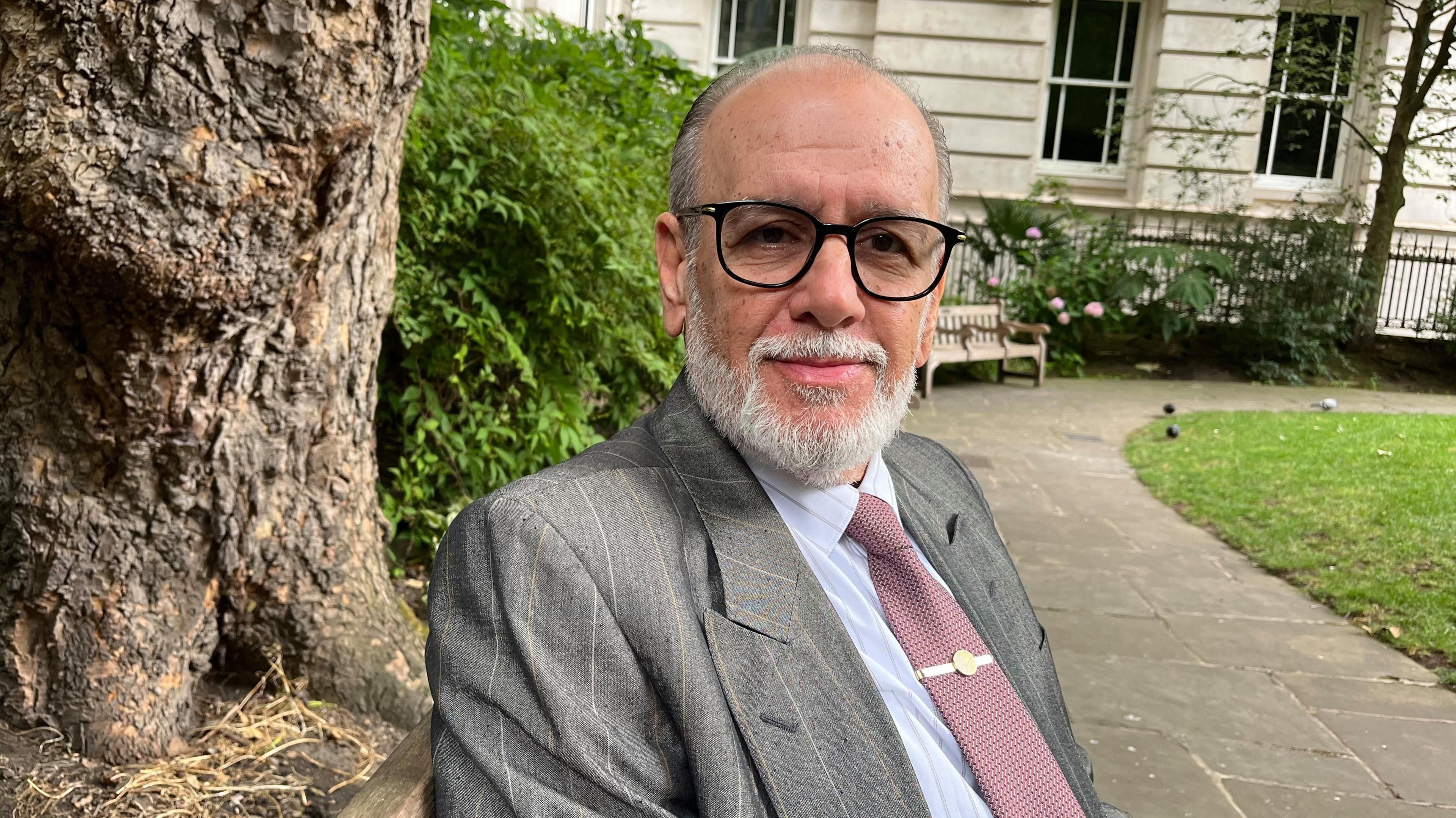 George Psaradakis in a mauve tie, grey suit and black glasses sits on a bench in a park with a tree trunk behind him.