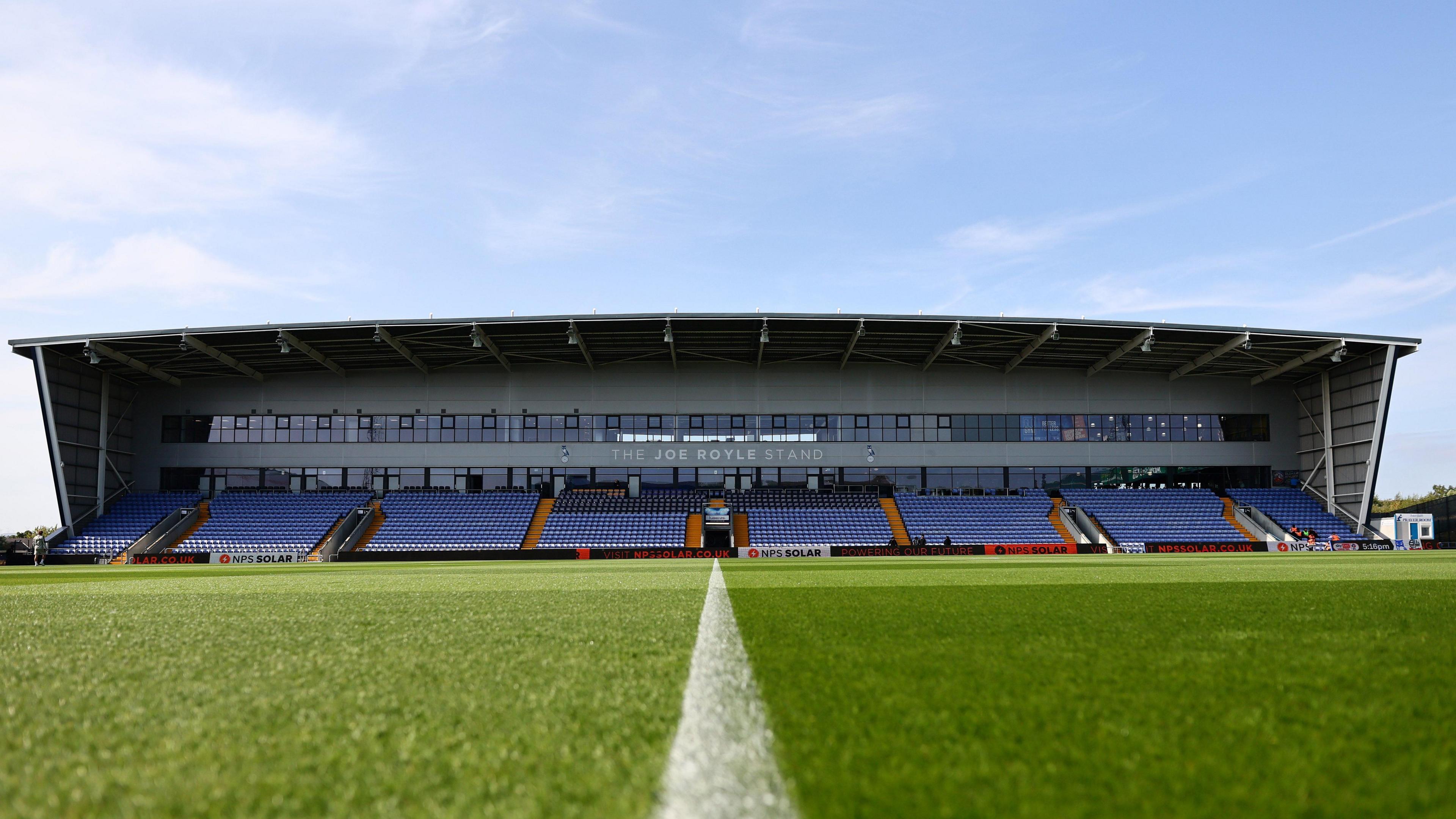 A ground-level view of the Joe Royle Stand at Oldham's Boundary Park on a sunlit early evening.