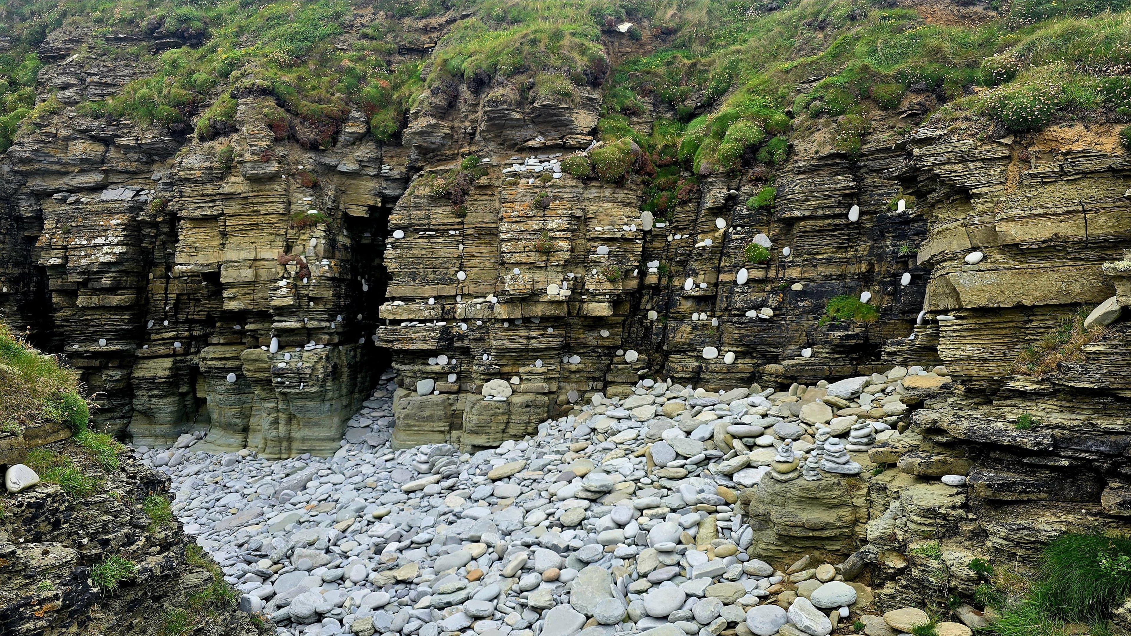 A dak sea wall with brown coloured crags with green moss on top. Below it is a collection of whte stones of different sizes and shades.