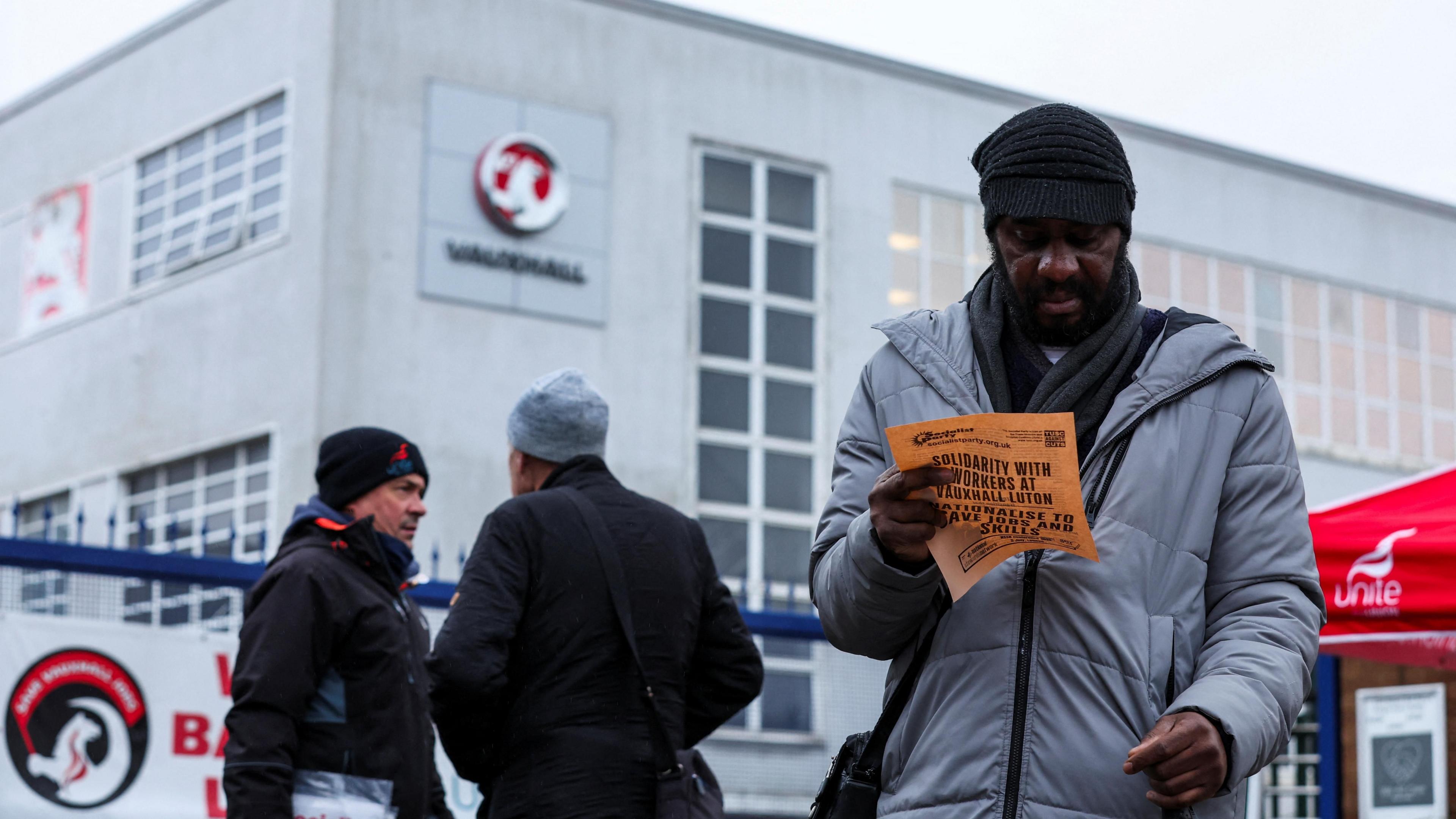 A man in a grey coat and a black hat is reading orange literature handed out by protesters outside the Vauxhall plant