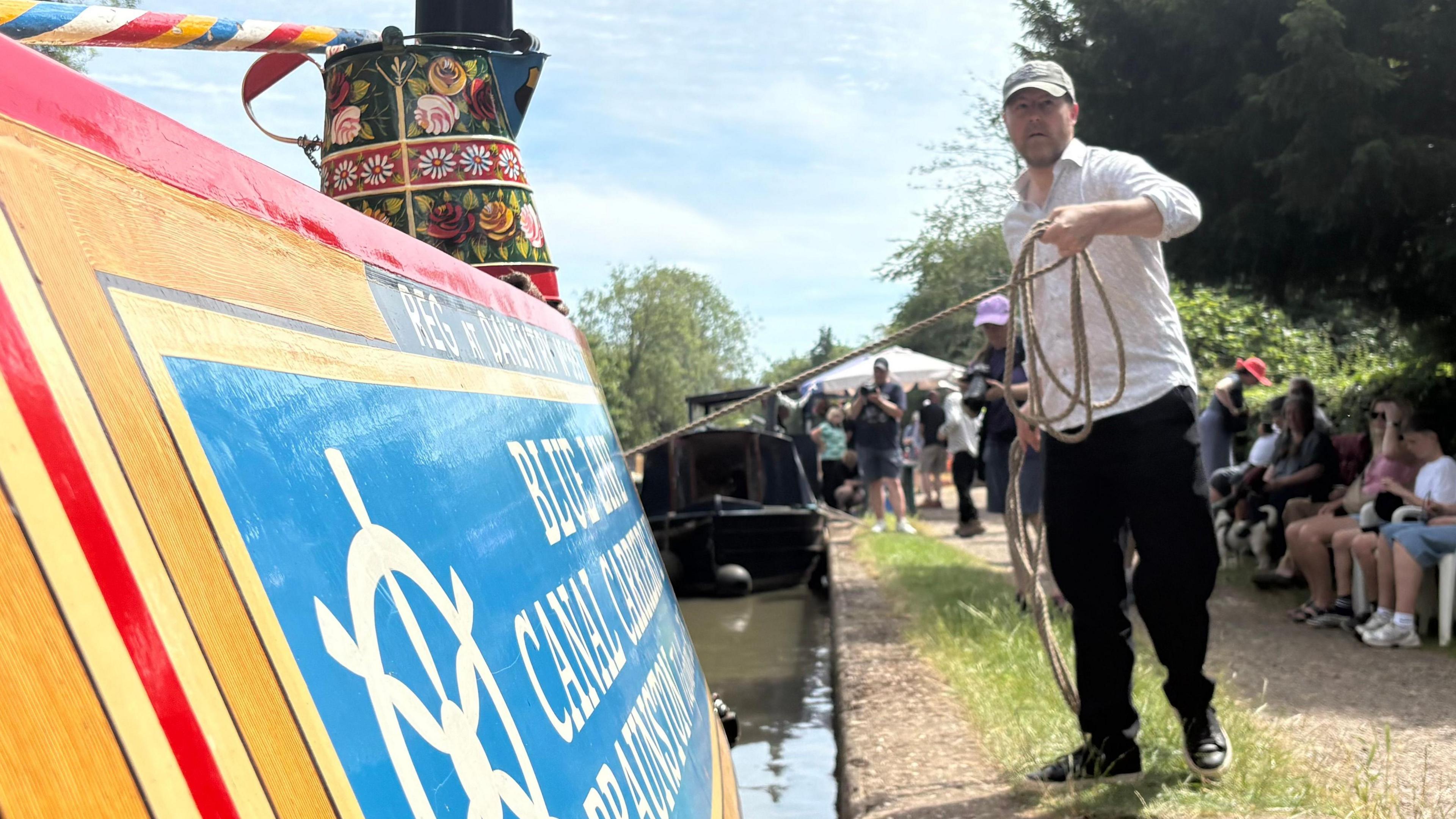 A brightly coloured canal narrowboat is moored alongside a path on a sunny day. The boat’s side, painted in red, yellow, and blue with decorative detailing, fills the left foreground. A tall, black chimney with gold bands and floral artwork rises from the boat, and part of a sign on the boat reads “B… Canal Cruis…” (partially obscured). On the right side of the image, Samuel West is standing on the towpath, holding a coiled rope that extends toward the boat. He is wearing a light-coloured shirt, dark trousers, and a cap, and is positioned slightly mid-step as if handling or pulling the rope.