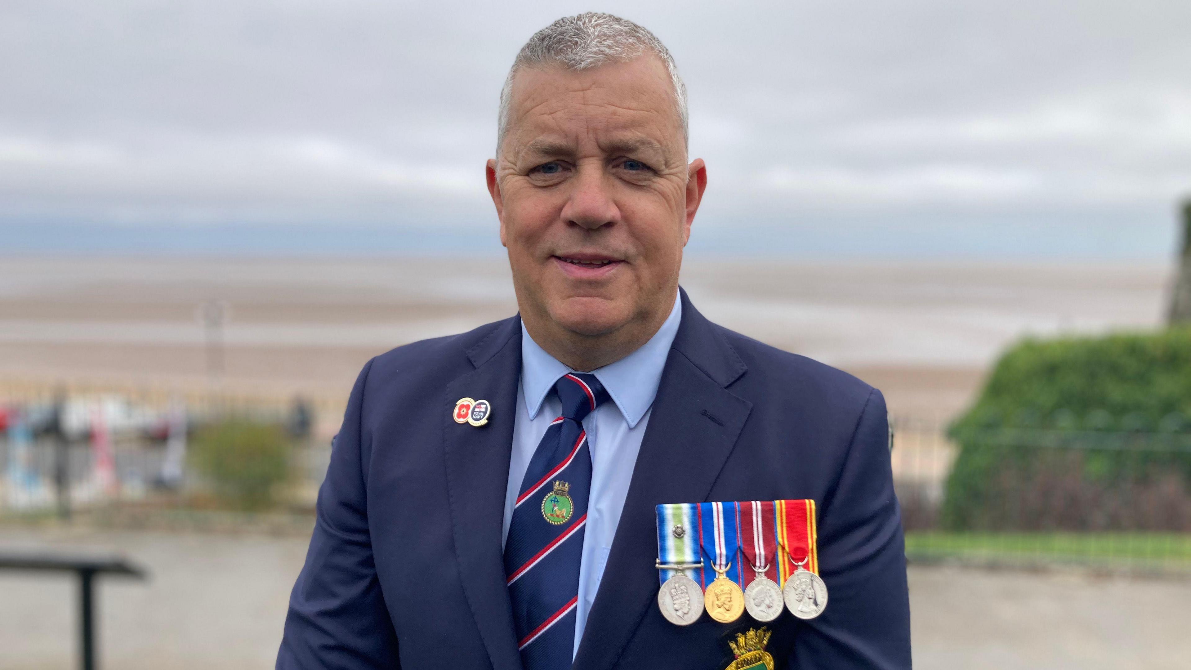 Nigel wears a smart dark blue suit with four medals on and is standing on the seafront, smiling at the camera.