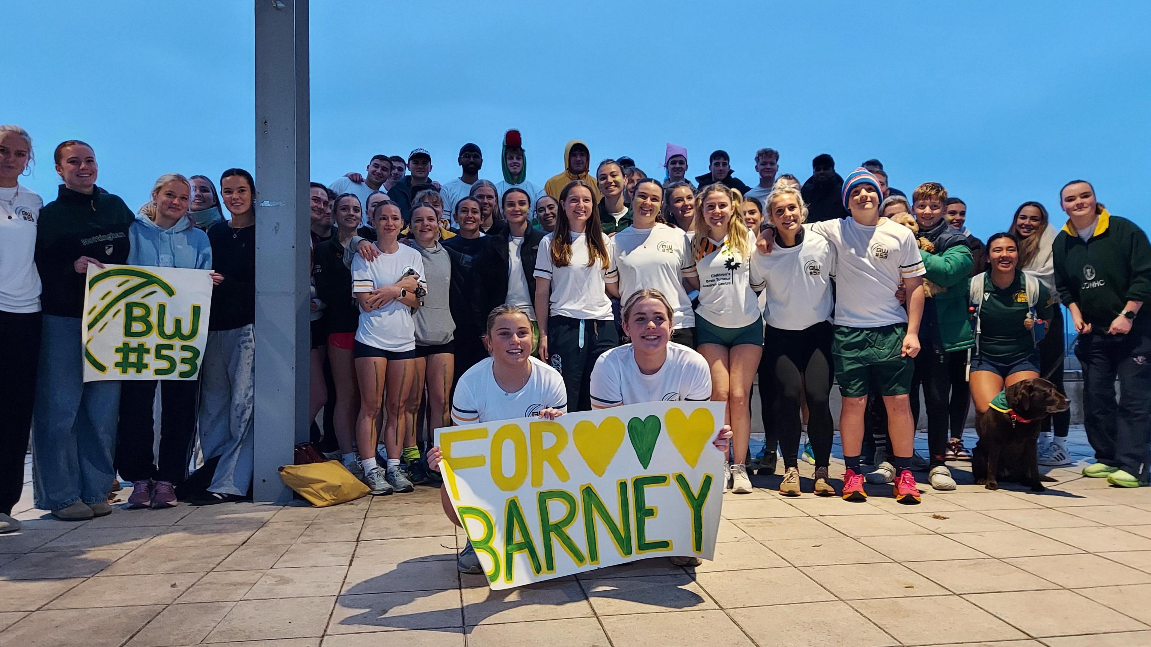 Friends, Abi Brant and Freya Tyler crouching down in front of a crowd of friends and family holding up a sign that reads "For Barney" with three hearts painted green and yellow. 