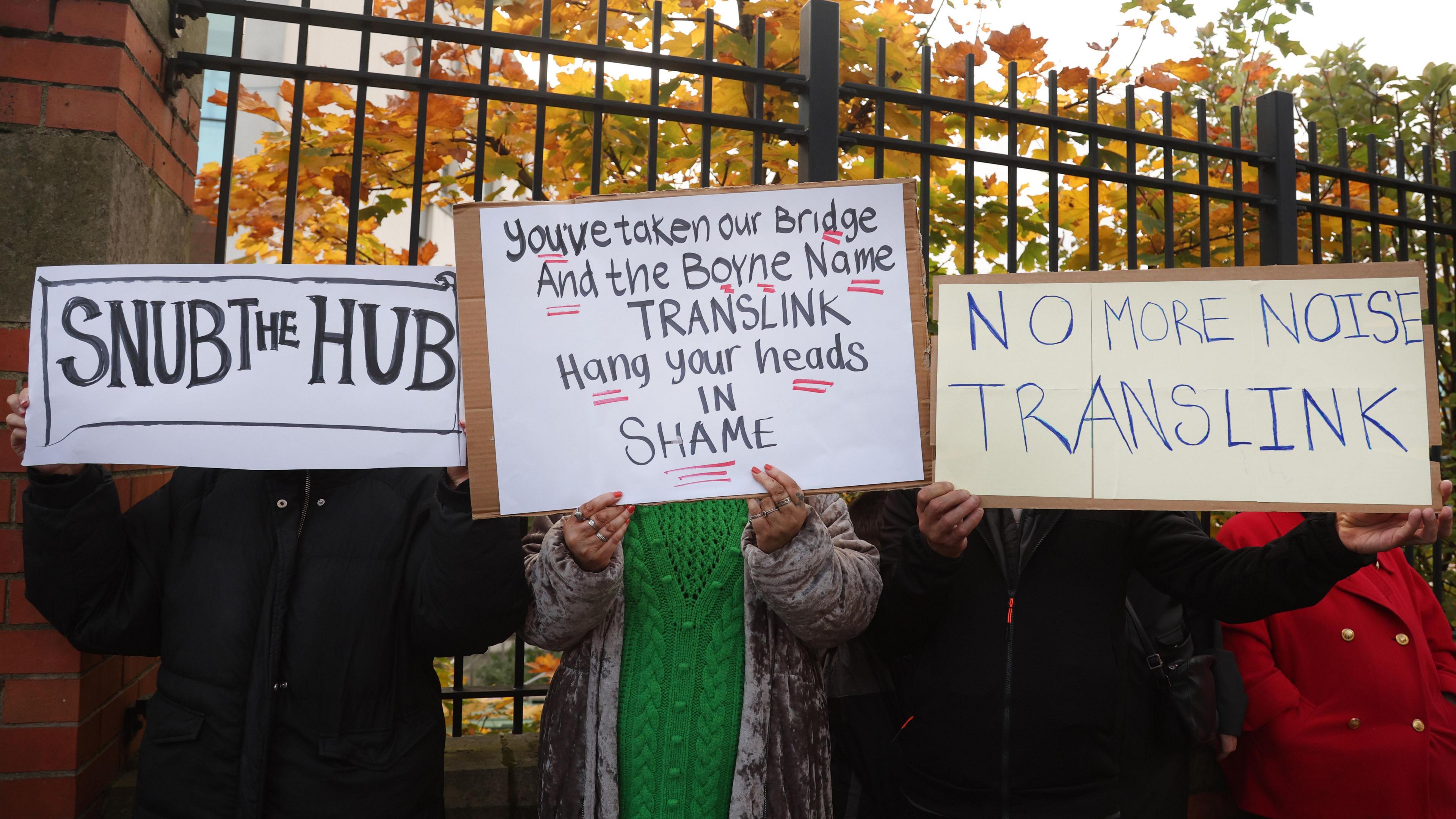 Three protestors on the Boyne Bridge. They are each holding a sign that covers their faces. On the left, one says 'Snub the Hub', the middle says 'You've taken our bridge and the Boyne Name Translink hang your heads in shame' and on the right the sign reads 'No more noise Translink'