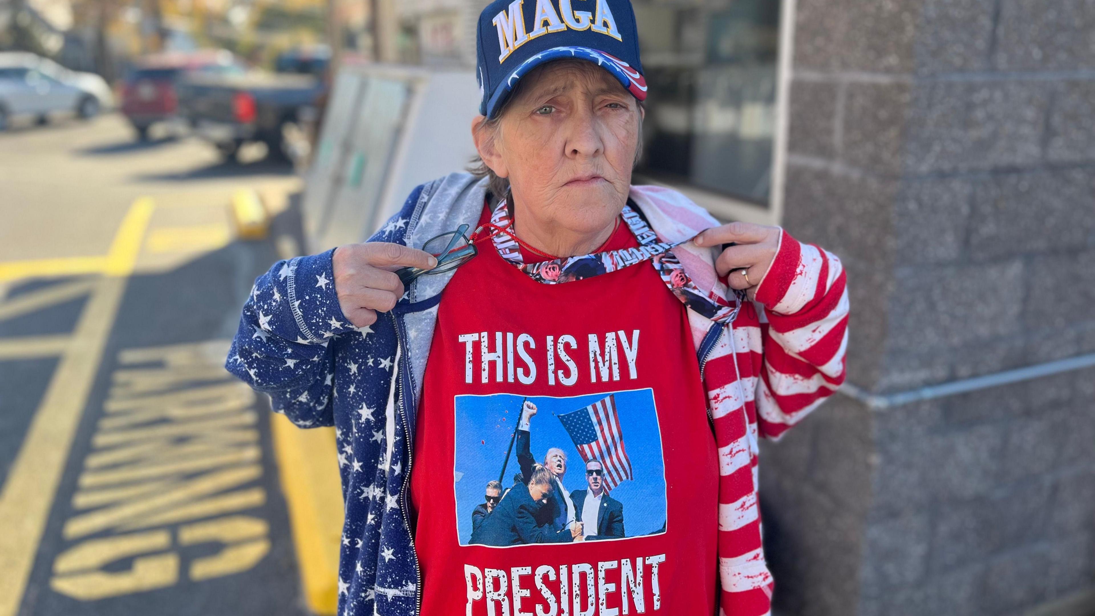 Patty is wearing an American flag patterned cap and jacket and a t-shirt showing Donald Trump, after he was shot on the ear, pump the air with his fist. 