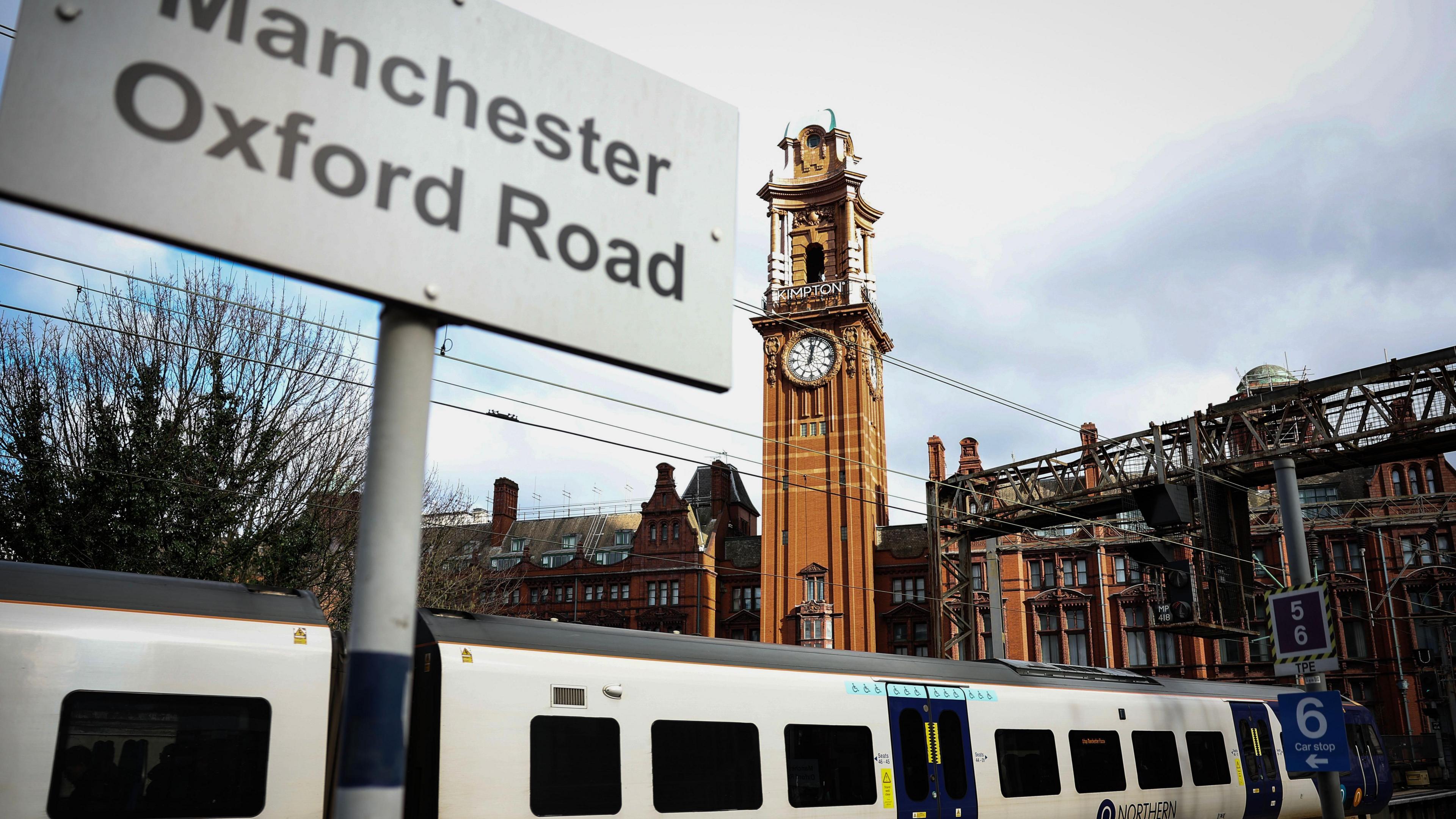 Sign saying 'Manchester Oxford Road' in front of train and tall, narrow clock tower at Manchester Oxford Road station.