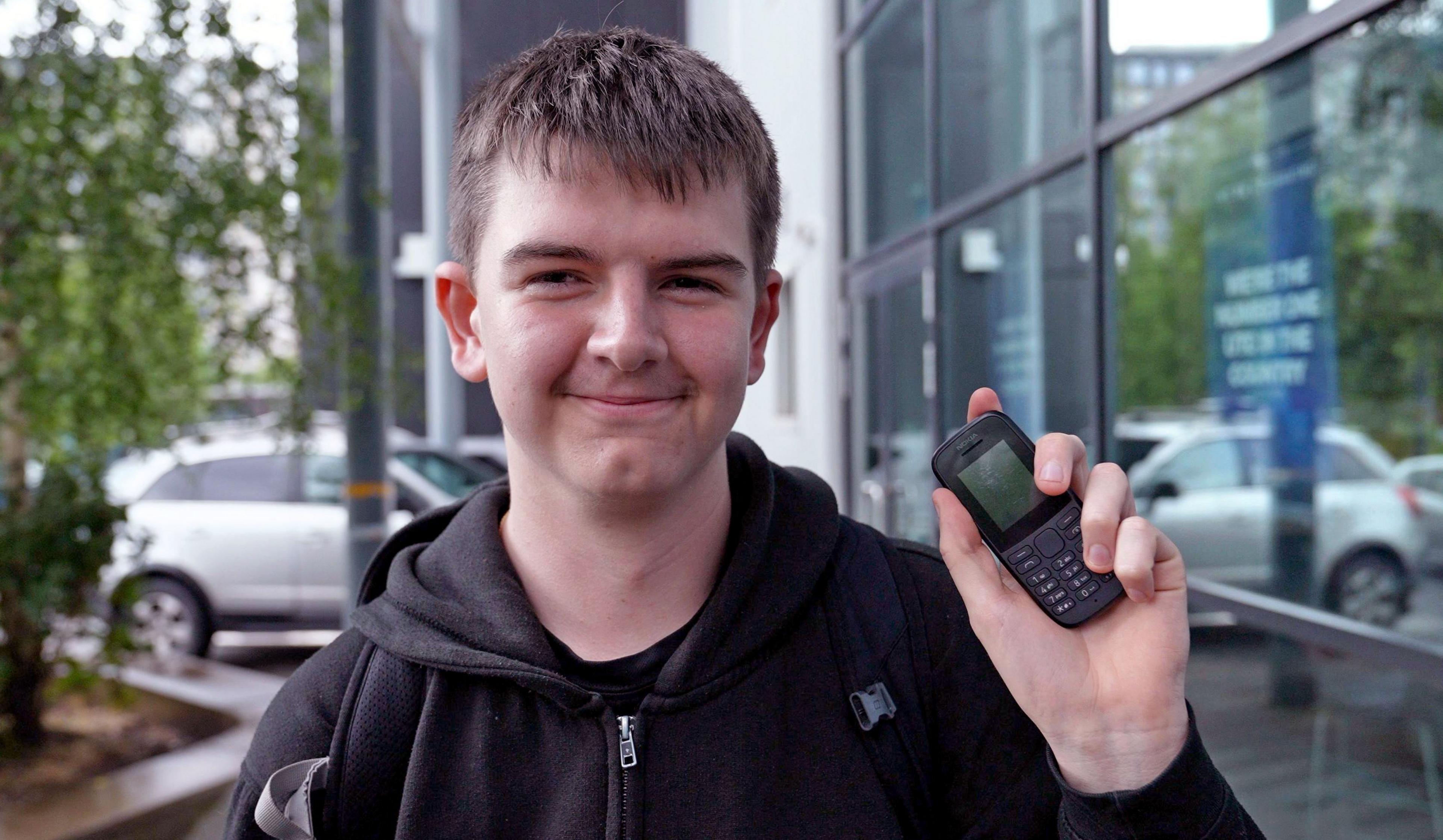 Will, 15, wears a black hoodie and smiles at the camera as he holds up his basic Nokia phone