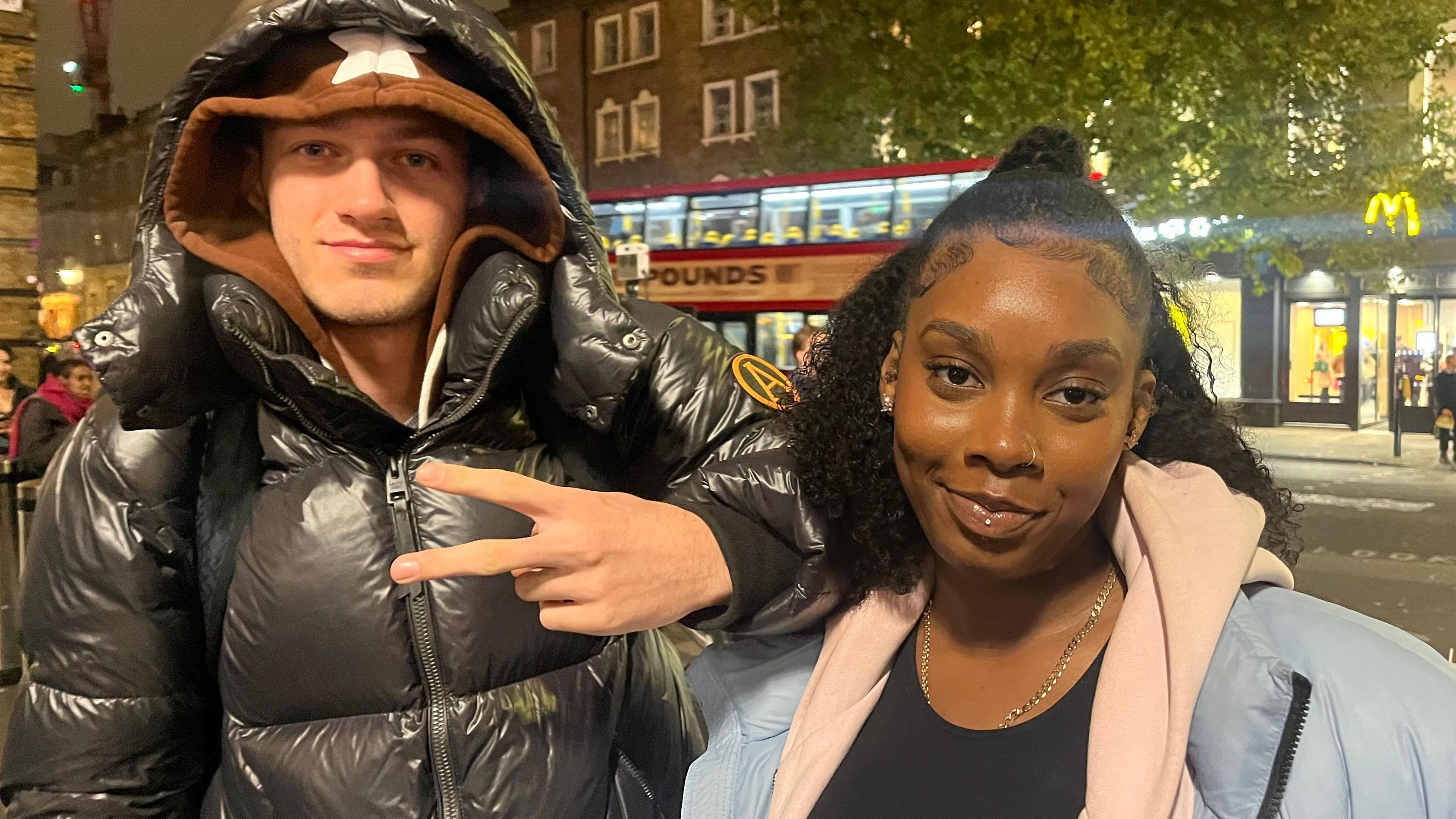 Louis is on the left wearing a brown hoodie covered by a black puffer jacket putting his fingers up in a v sign. Kaianne is on the right wearing a black top with a pink hoodie and a blue puffer - smiling into the camera.