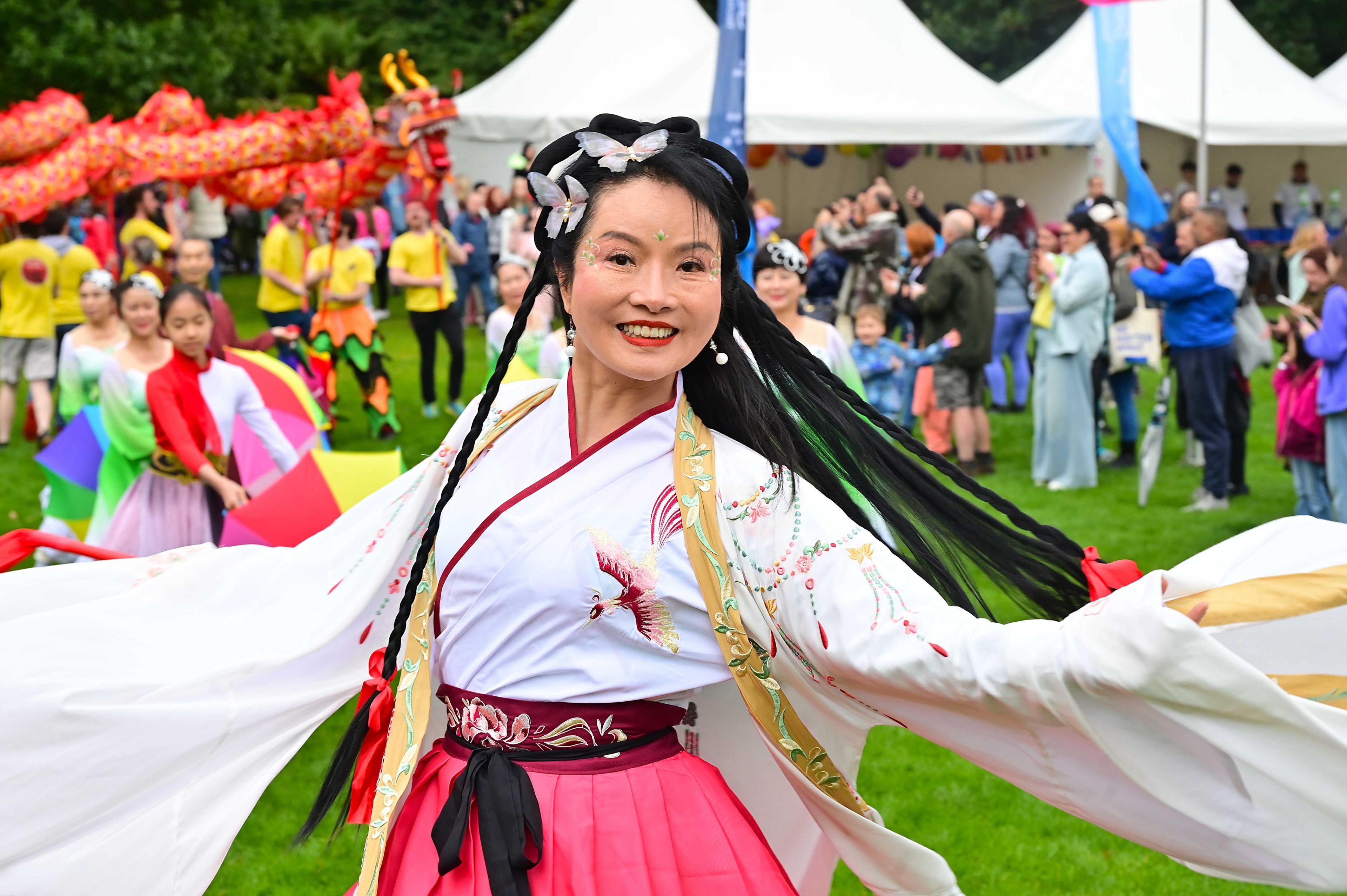 A woman smiling in a white, long sleeved top with birds embroidered and a pink skirt, tied at waist with black bow. Stood in front of a large, colourful crowd at Mela Day with white tents in the background.