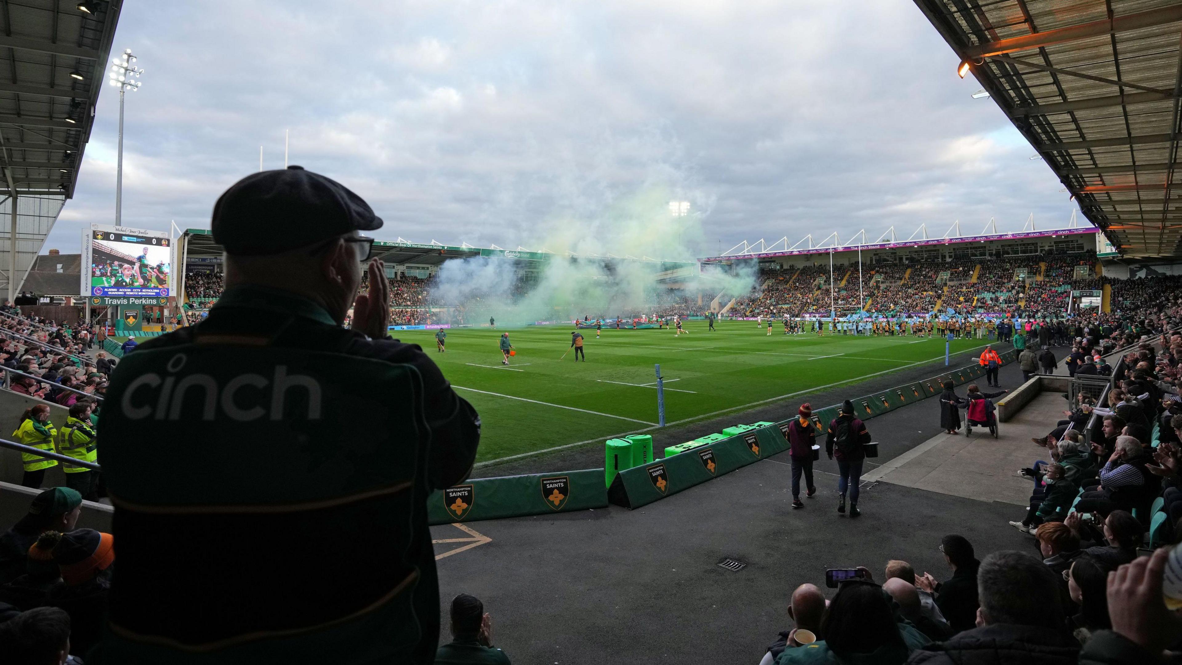 A general view of Franklins Gardens, a purpose-built rugby stadium in Northampton. There are players on the pitch with many supporters in the surrounding stands. There is a close-up shot of a man's back who is wearing a cap. There are fireworks going off on the pitch.