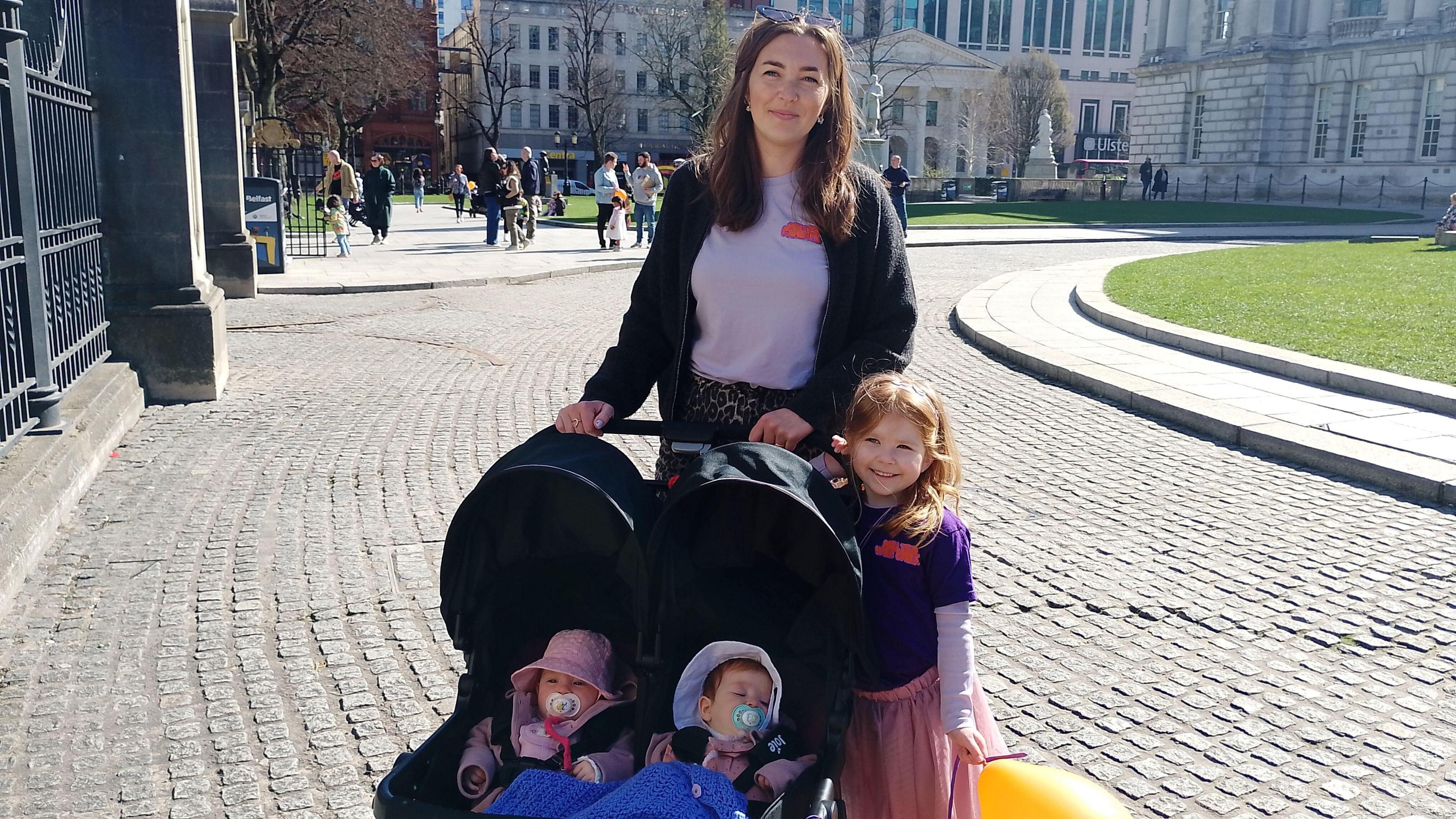 A woman, with brown hair, wearing a white top and black jacket, standing with a double pram with two babies in it and a child with red hair, wearing a purple t-shirt and pink skirt. They are standing in the grounds of Belfast City Hall.