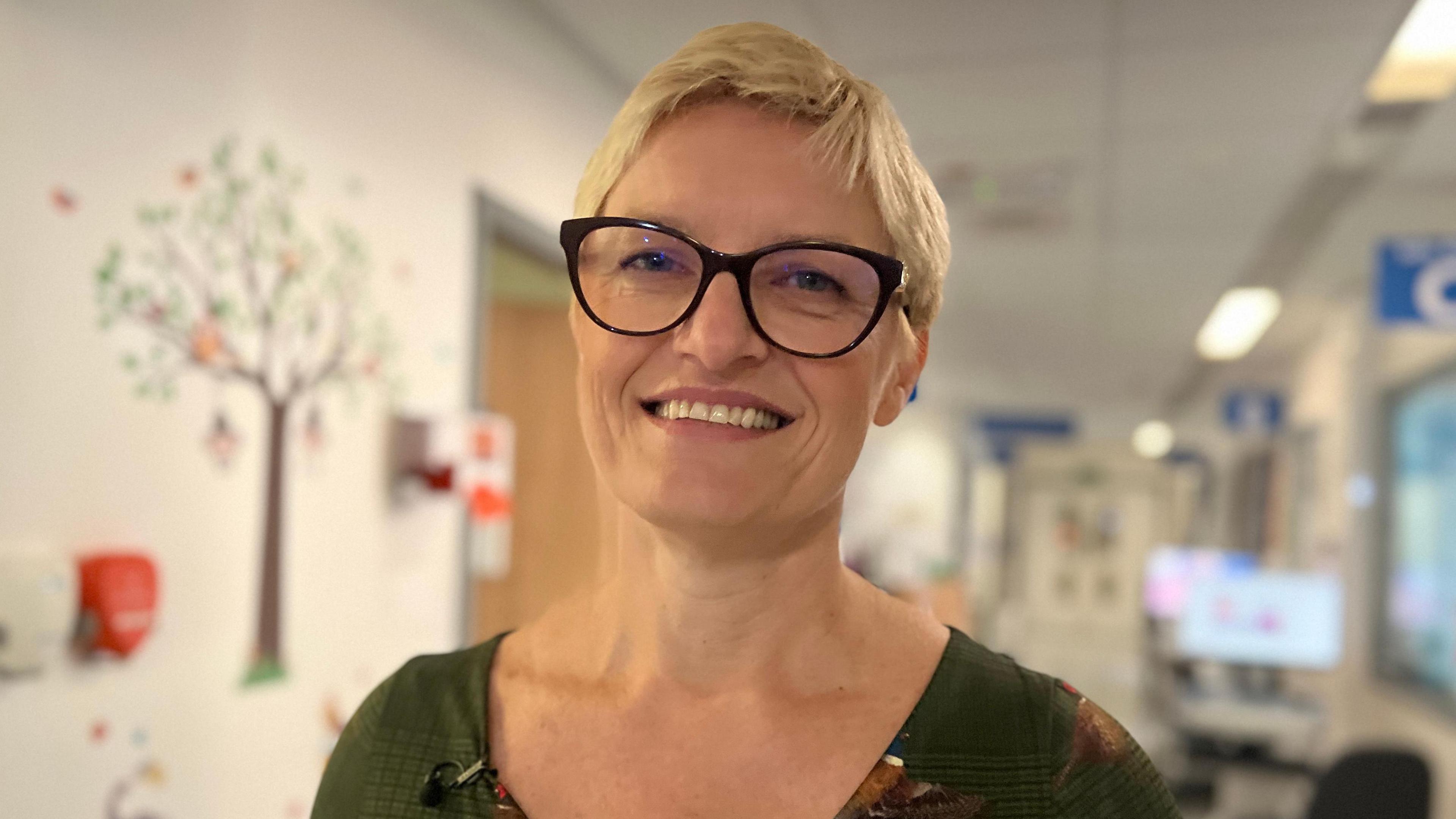 Dr Katarzyna Gajewka-Knapik, who has short blonde hair and wears large black framed glasses and a green top. She is smiling towards the camera and behind her is a hospital corridor.