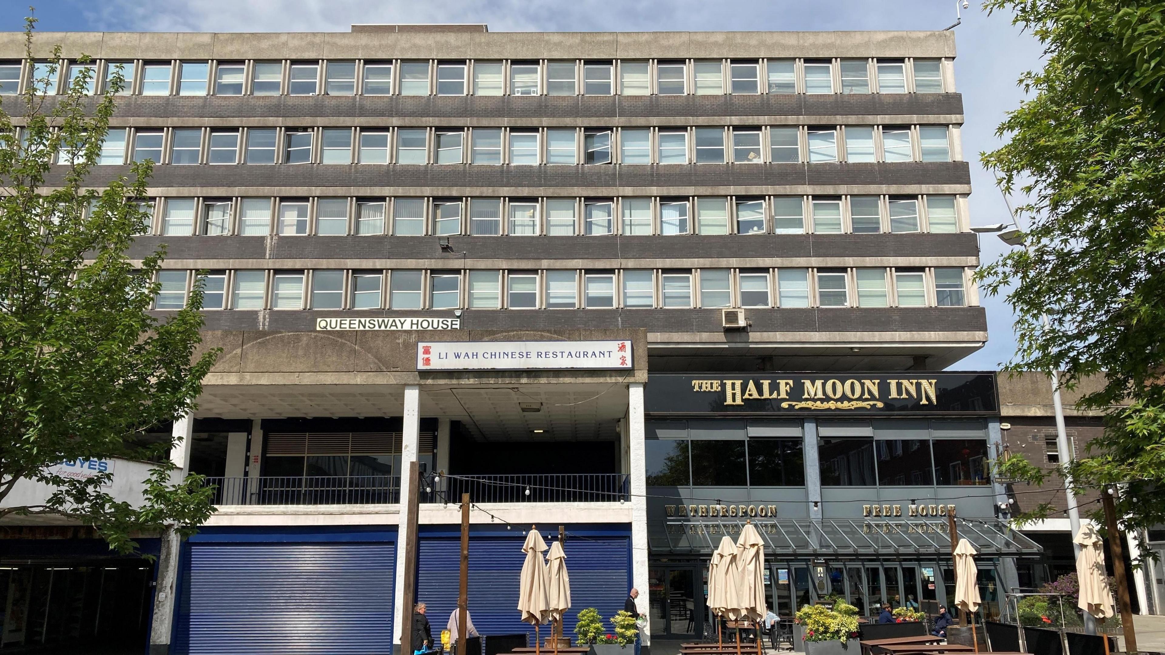 West Precinct is a large, concrete Brutalist-style building. Pictured are two businesses at the base of the block - the Half Moon Inn and a Chinese restaurant with its blue shutters down.