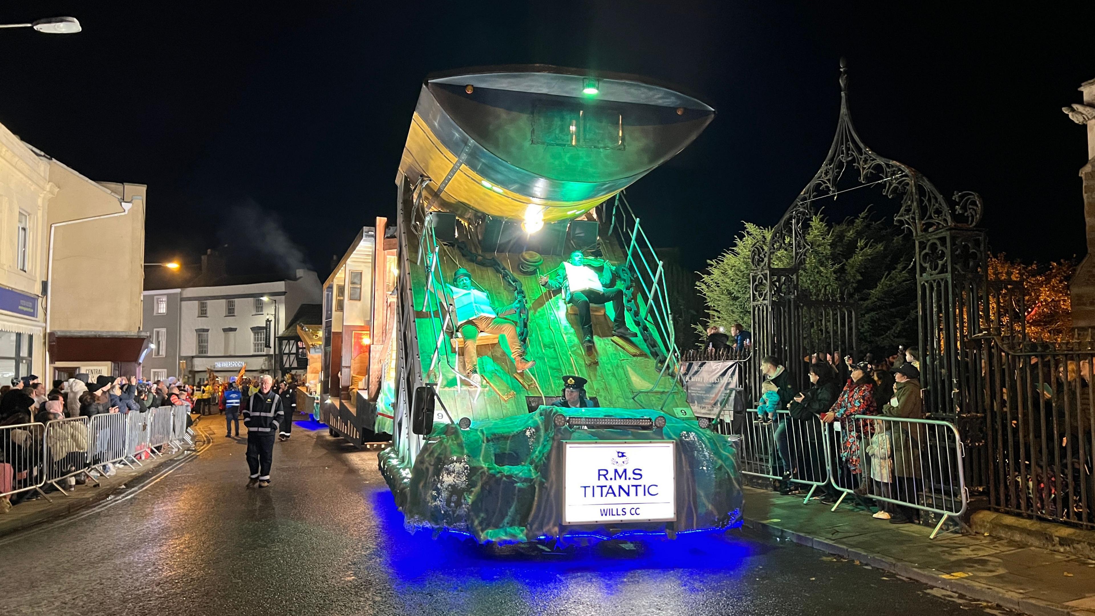 A Titanic-themed cart is being driven down a road in Bridgwater. The car has been designed to look like the sinking Titanic ship. The front of the cart is sloping, with two people appearing to slide down the wooden floor of the boat. There is a sign at the front of the cart that says R.M.S Titanic. There are dozens of spectators lining the streets behind metal barriers.