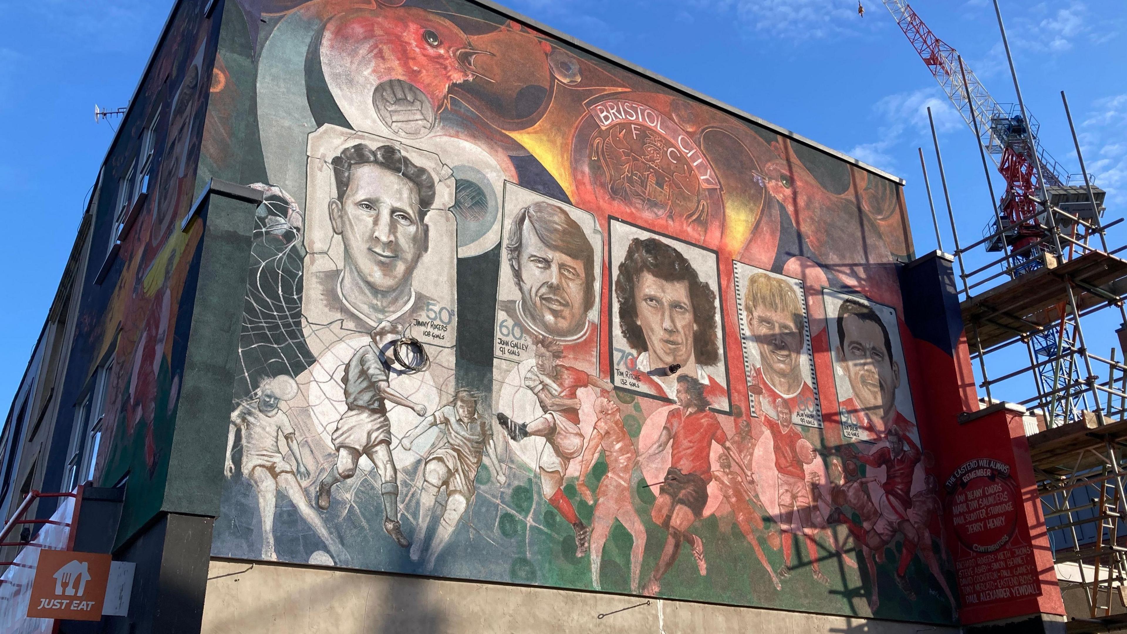 The mural, pictured from street level, which is on the first floor of a building. It shows the faces of five footballers, on what appear to be vintage cards. Below the cards are images of footballers playing in a variety of poses, while above the cards is a version of Bristol City's emblem and two robins - the symbol of the club.