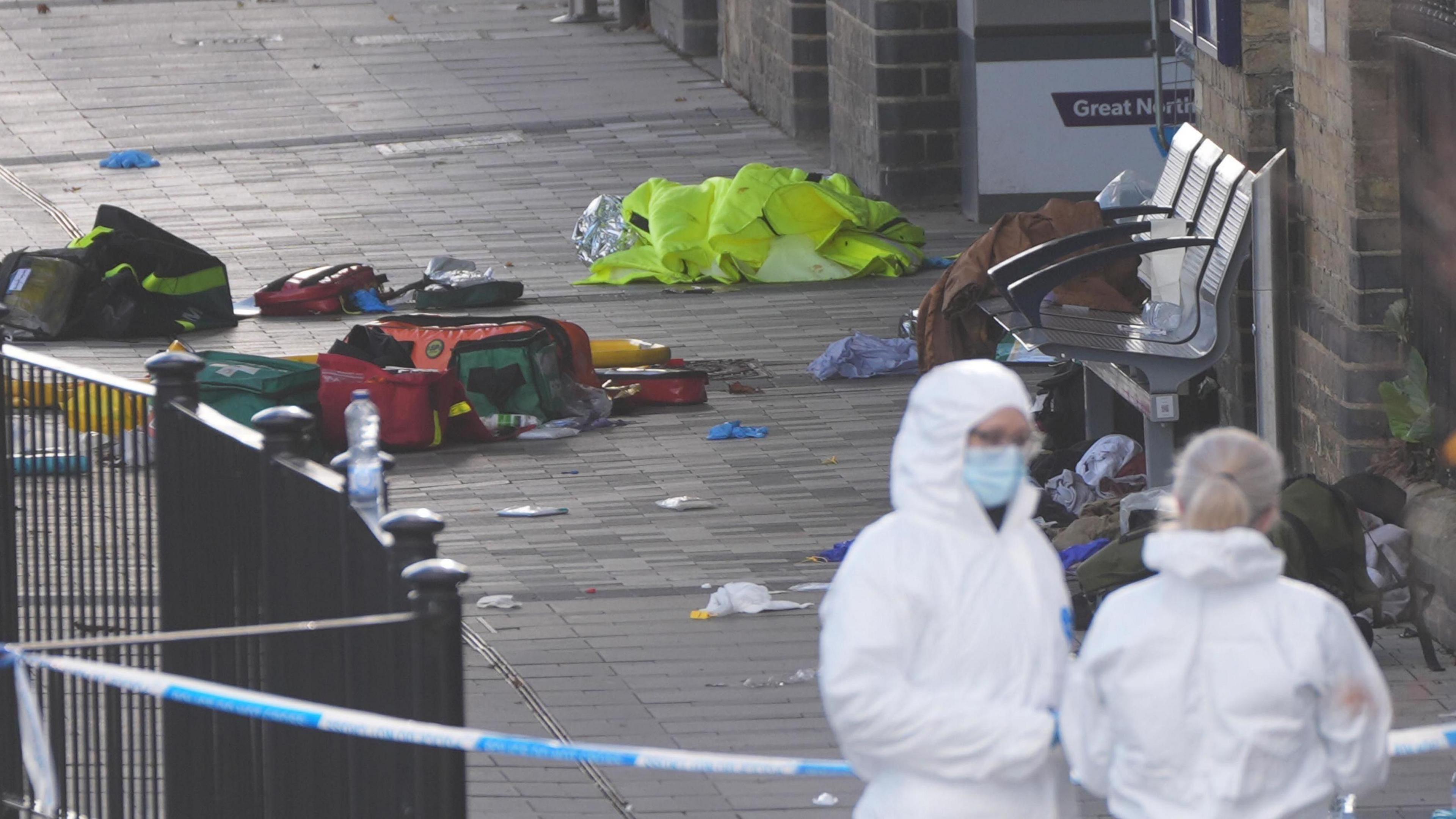 Two people are stood on a train platform and are wearing white safety suits. They are stood behind blue and white police tape which has cordoned off an area which is full of rubbish and ambulance equipment. 